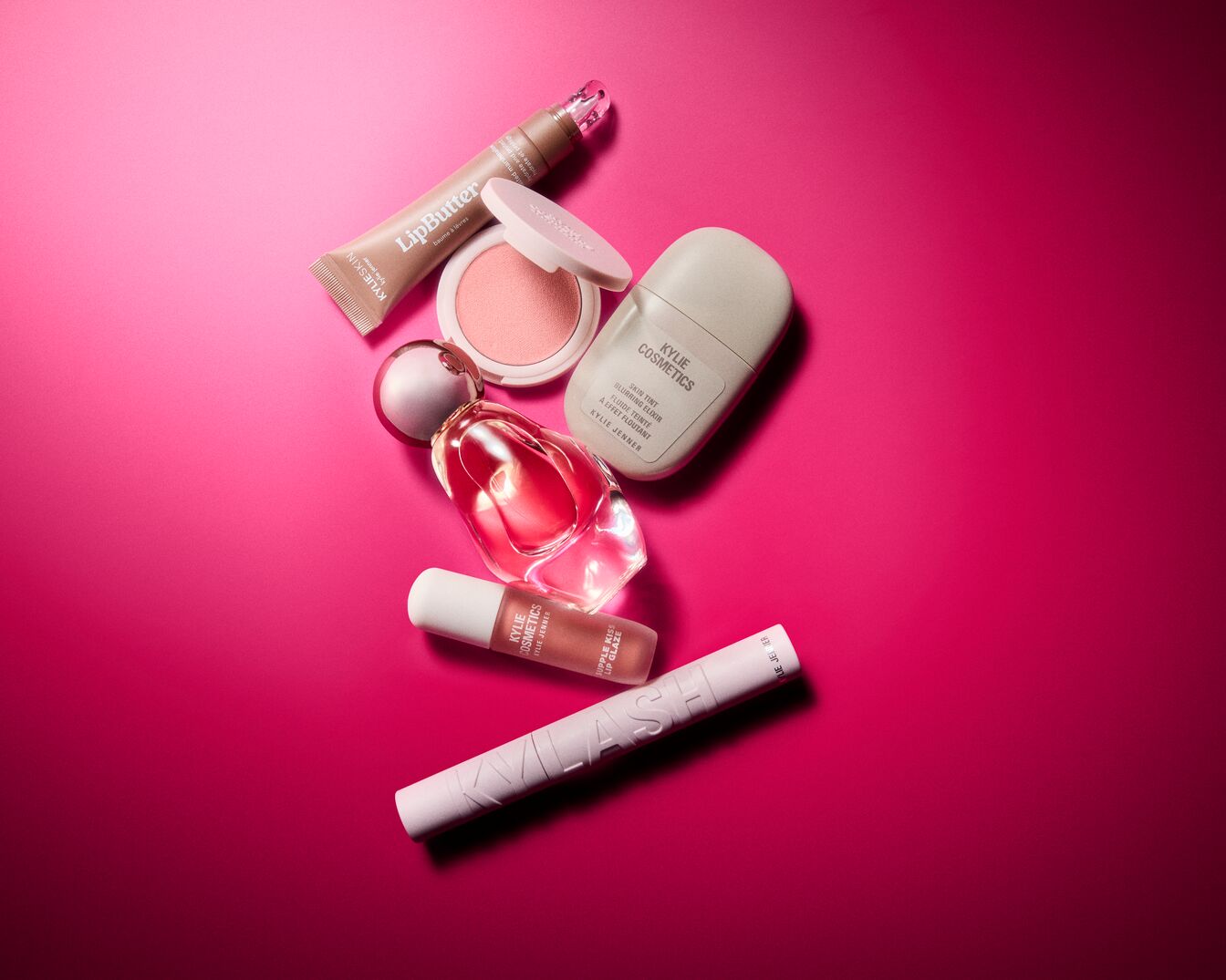 Ant ryškiai rožinio fono išdėstytas „Kylie Cosmetics“ makiažo priemonių rinkinys, kurį sudaro švelnių atspalvių lūpų blizgiai, kompaktiniai skaistalai, skystas odos tonavimo fluidas, kvepalų flakonas ir blakstienų tušas.