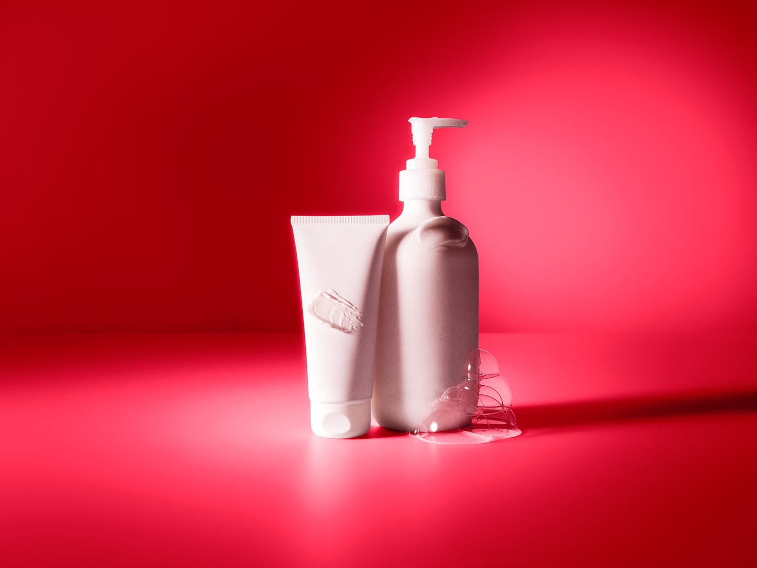 Een witte tube en een witte pompflacon staan op een rode ondergrond, met een veeg crème op de tube en zeepbellen bij de flacon, wat de textuur en het gebruik van de producten benadrukt.