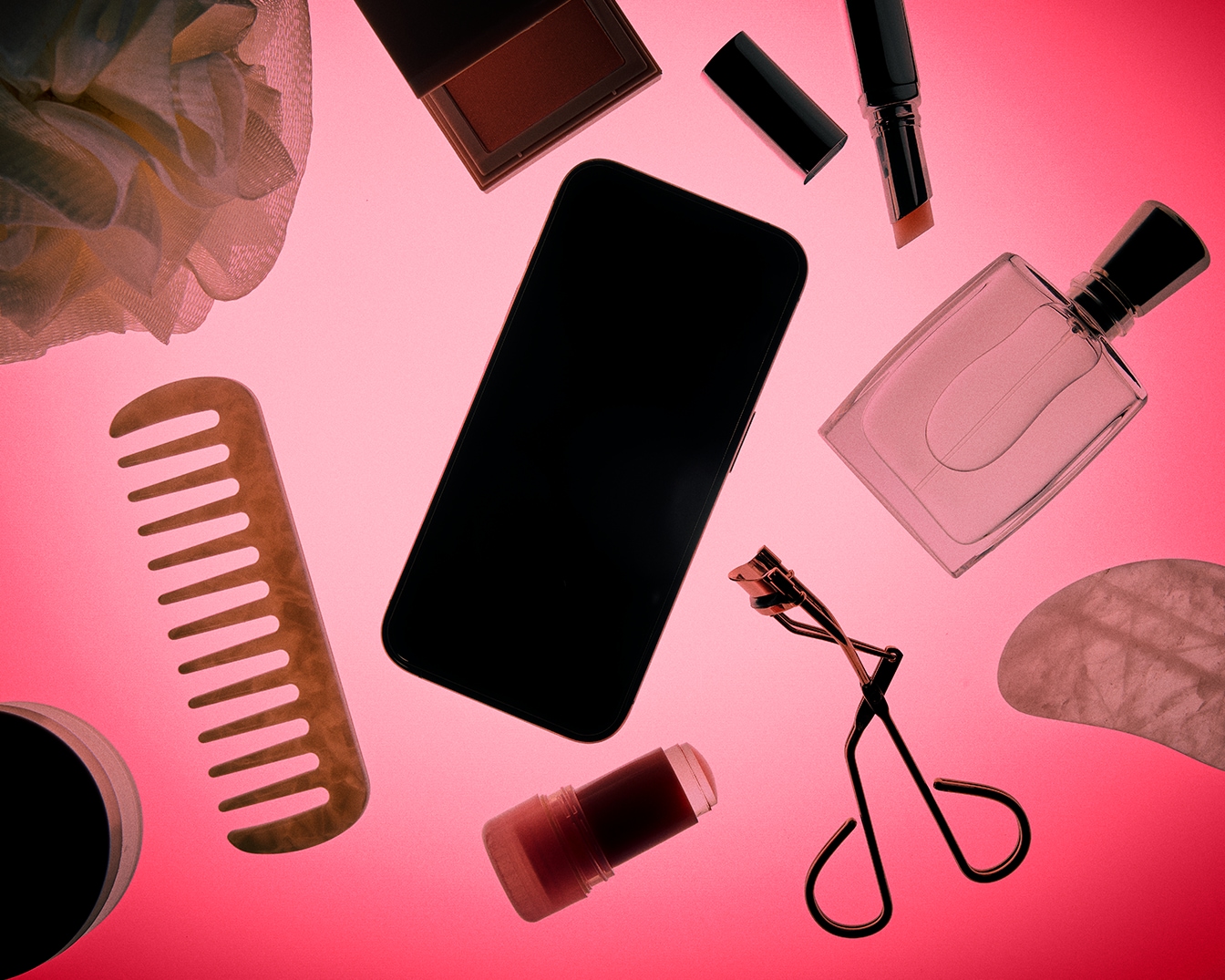 Een verzameling van beautyproducten en accessoires, waaronder een mobiele telefoon, parfum, make-up, een kam, een wimperkruller en een badspons, allemaal geplaatst op een levendige roze achtergrond.