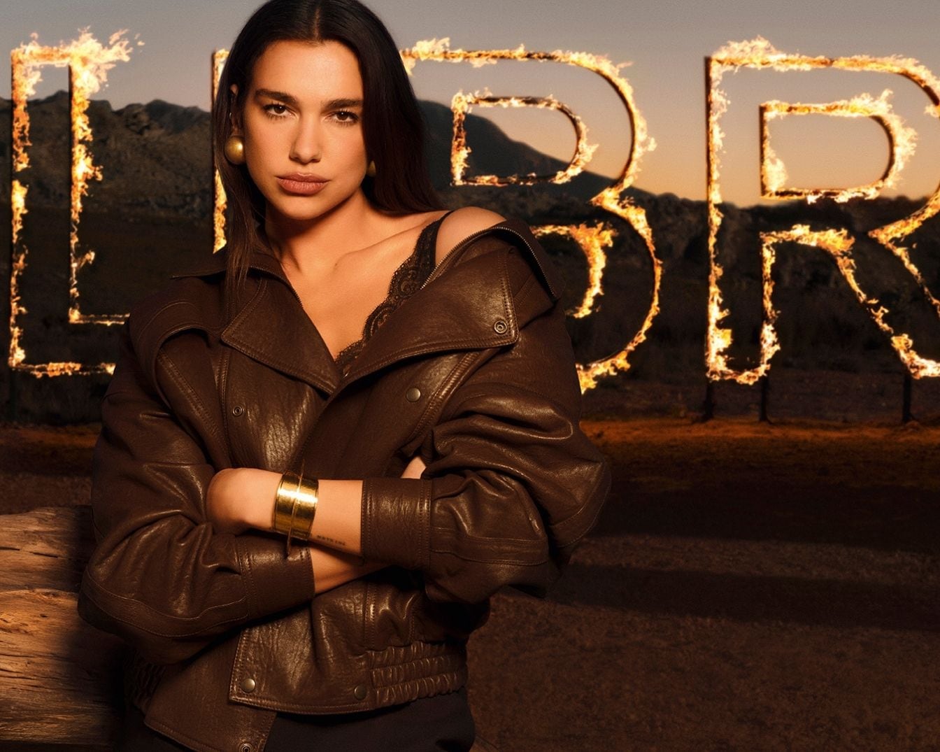 Dua Lipa barna bőrkabátban és fekete csipkés felsőben pózol, arany fülbevalóval és karkötővel, a háttérben lángoló betűkkel, amelyek a "LIBRE" szót formázzák.