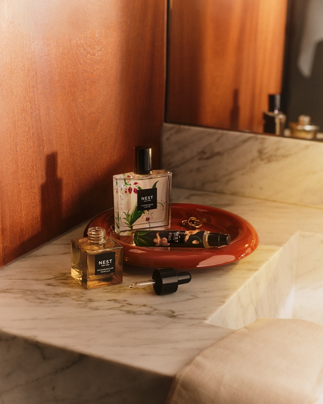 Ant marmurinio stalviršio vonios kambaryje stovi trys „Nest New York“ produktai: „Cardamom Latte“ kvepalai, „Madagascar Vanilla“ aliejus ir „Indian Jasmine“ rankų kremas, išdėstyti ant raudono padėklo su auksiniais žiedais.