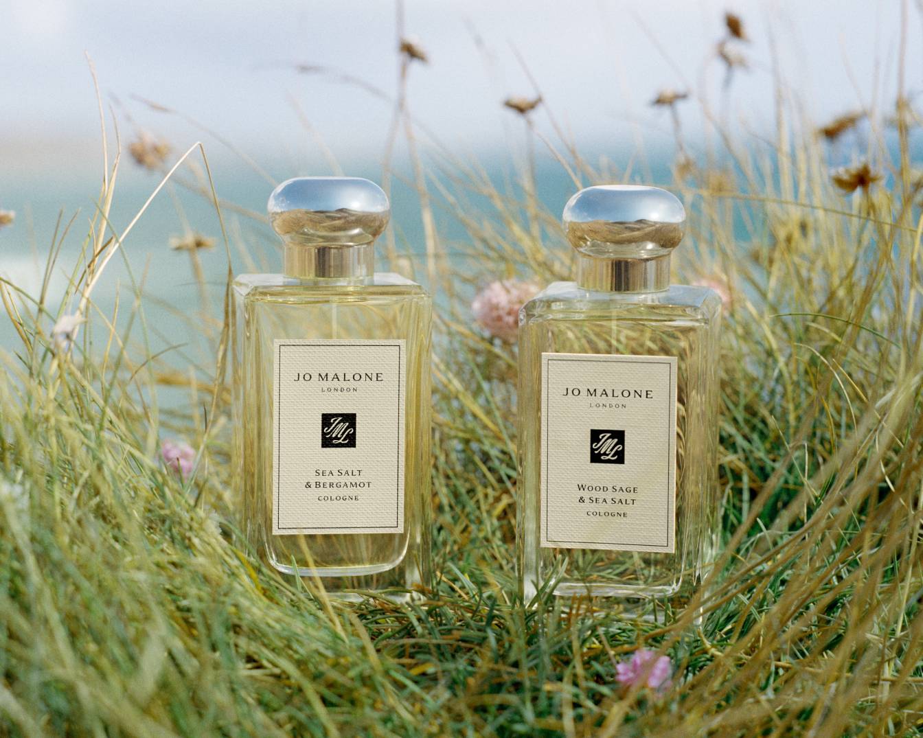Dwa flakony perfum Jo Malone London, jeden Sea Salt & Bergamote, drugi Wood Sage & Sea Salt, stoją w wysokiej trawie na tle oceanu.