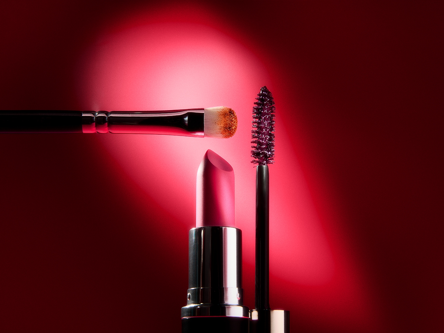 Un pinceau de maquillage, un rouge à lèvres rose vif et une brosse à mascara violette sont élégamment disposés sur un fond rouge éclatant, créant une ambiance glamour.