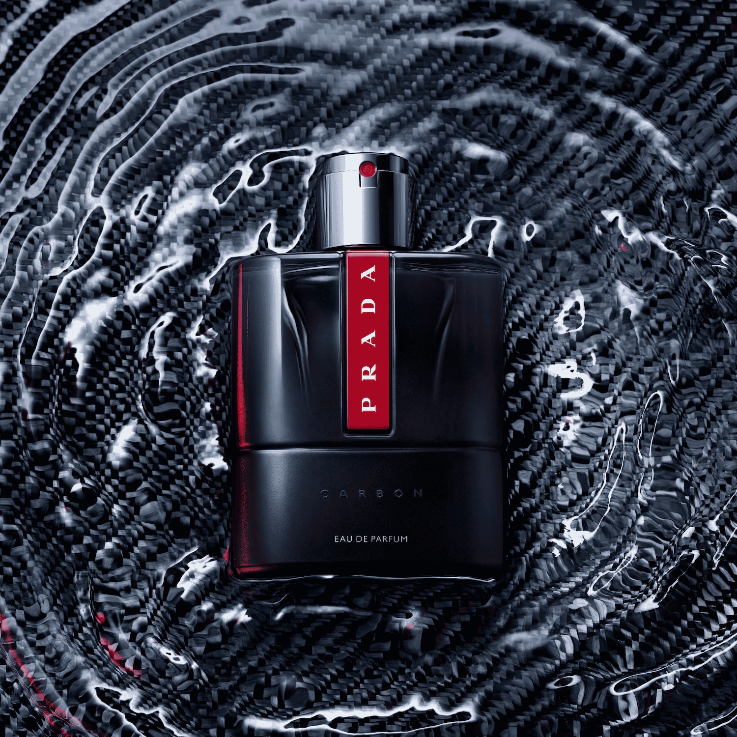 Prada Carbon Eau de Parfum je prezentována v elegantní černé lahvičce s červeným pruhem a stříbrným rozprašovačem, obklopená vlnící se vodou na texturovaném pozadí.