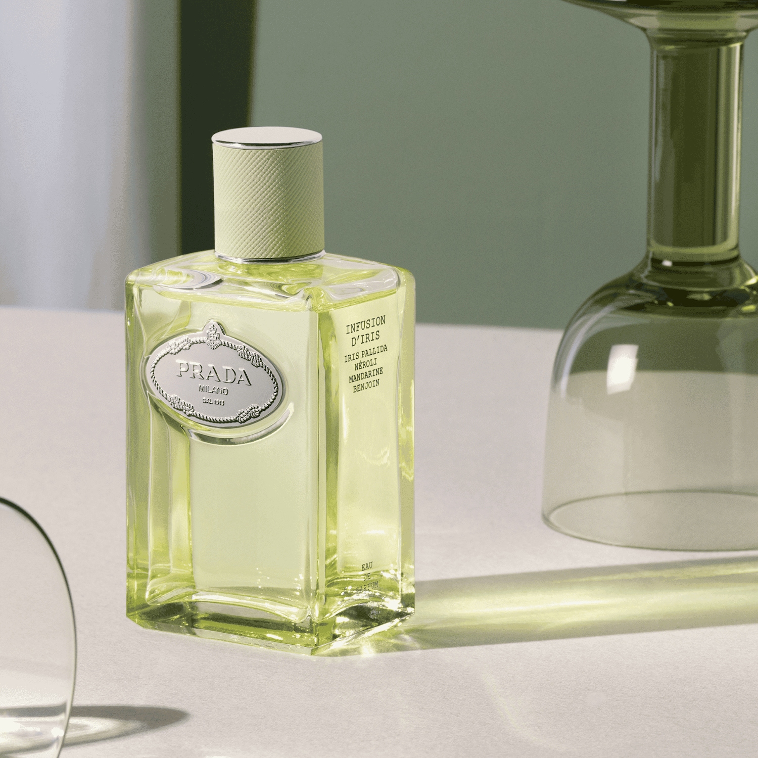 Prada Infusion d'Iris je elegantní parfémová voda ve čtvercové skleněné lahvičce s bledě zelenou tekutinou a stříbrným oválným štítkem s nápisem Prada Milano, doplněná světle zeleným víčkem s texturou.