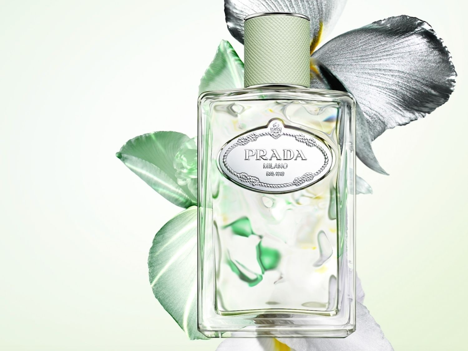 Prada Infusion d'Iris Eau de Parfum ve skleněném flakonu s jemně zeleným uzávěrem, obklopená zelenými a stříbrnými okvětními lístky, evokuje svěžest a eleganci.