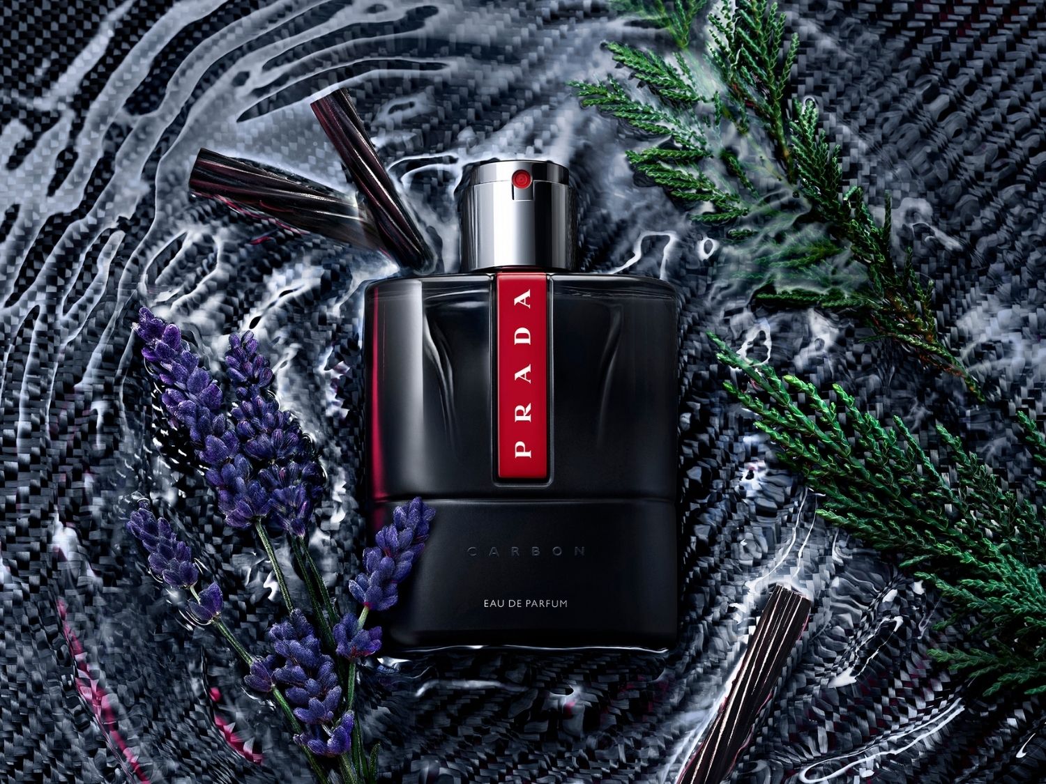 Prada Luna Rossa Carbon Eau de Parfum v čiernej fľaši s červeným logom Prada, obklopená levanduľou a zelenými vetvičkami na tmavom, textúrovanom pozadí s vodnými vlnkami.