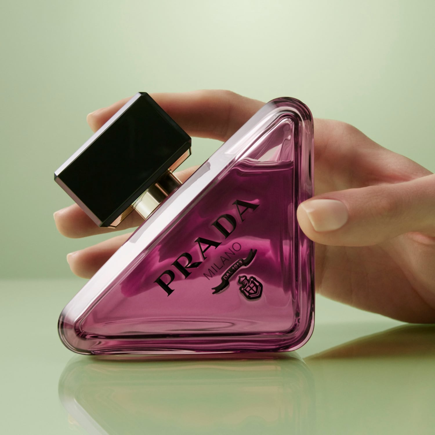Ruka drží fľaštičku parfumu Prada Paradoxe v tvare trojuholníka s ružovou tekutinou a čiernym vrchnákom, ktorá odráža svetlo na svetlozelenom pozadí.