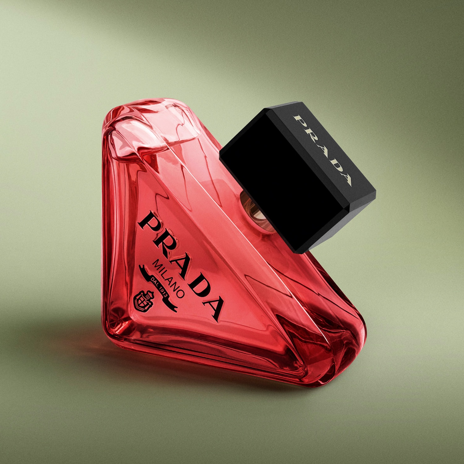 Prada Paradoxe, červený parfum v tvare trojuholníka s čiernym vrchnákom, leží na boku na zelenom pozadí.