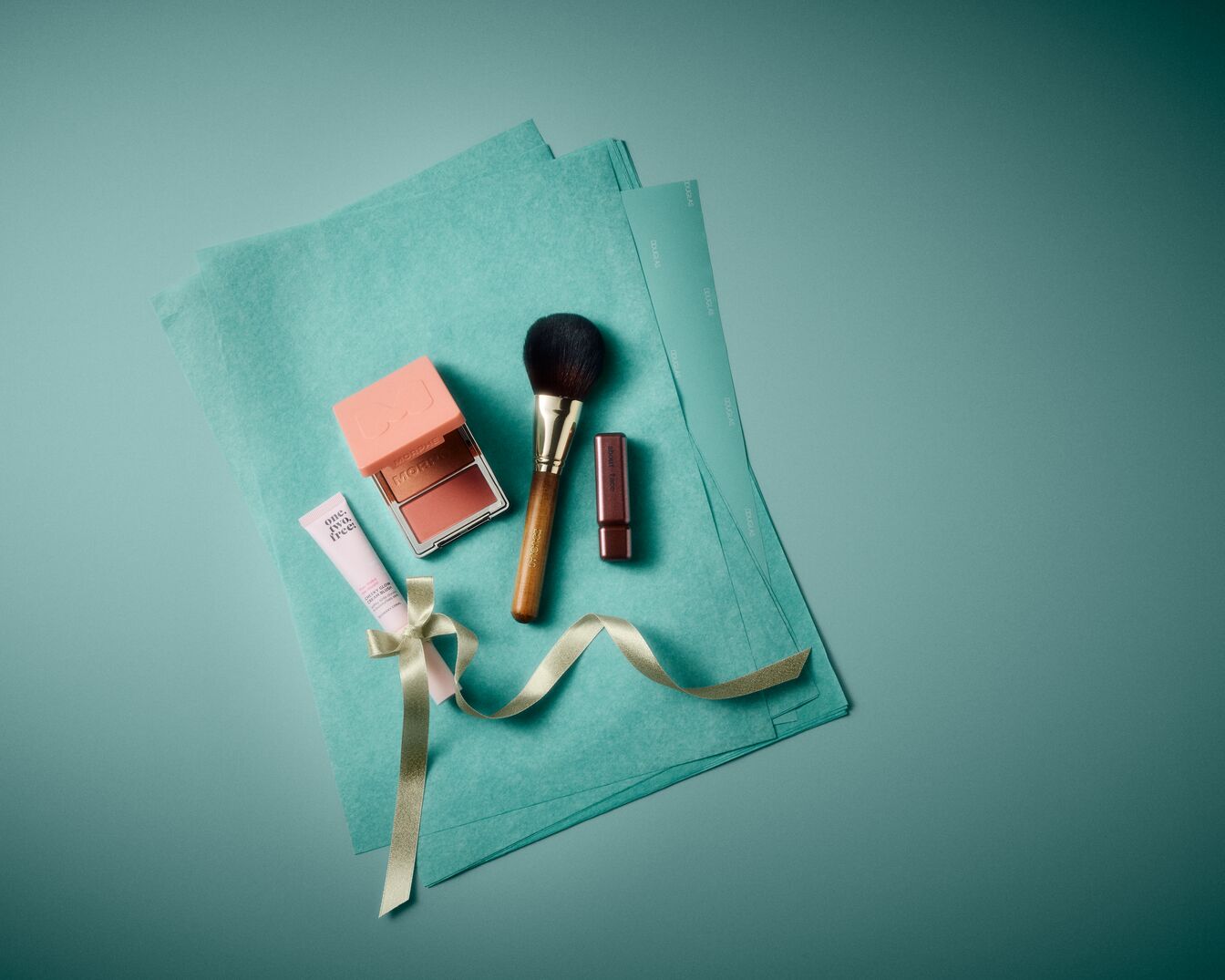 Een Morphe blush in een perzikkleurig doosje, een make-upkwast met houten handvat, een roze lippenstift en een lichtroze tube One.Two.Free. crème liggen op turkoois papier, versierd met een goudkleurig lint.