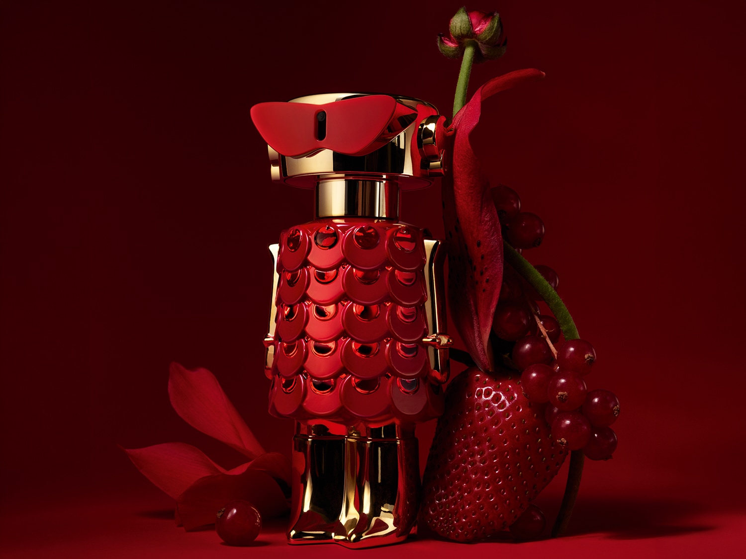Un flacone di profumo rosso e oro a forma di robot, con occhiali da sole rossi, è affiancato da un fiore rosso, fragole e ribes rosso su uno sfondo rosso.