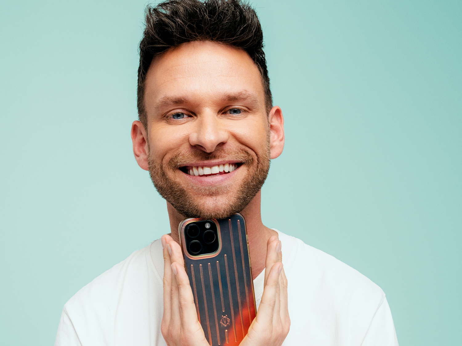 Een lachende man met een baard en blauwe ogen houdt een smartphone met een donkere, gestreepte hoes met oranje accenten voor zijn kin.