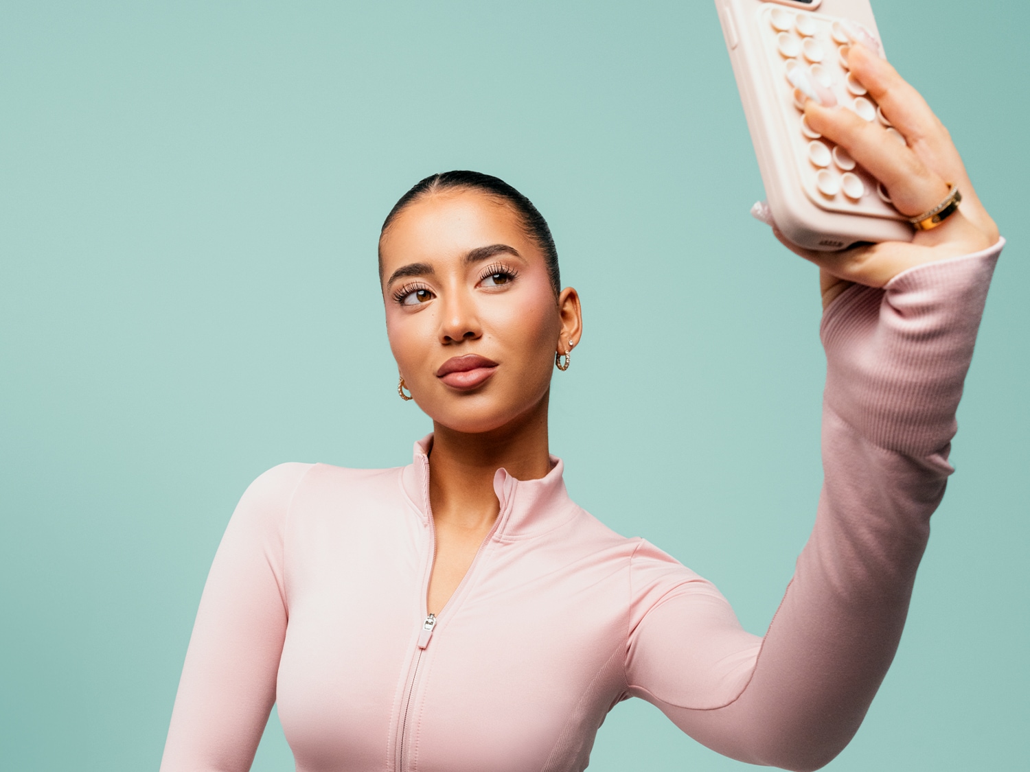 Een vrouw met een roze sporttop en een strakke paardenstaart maakt een selfie met een lichtroze telefoonhoesje, haar blik is naar links gericht.