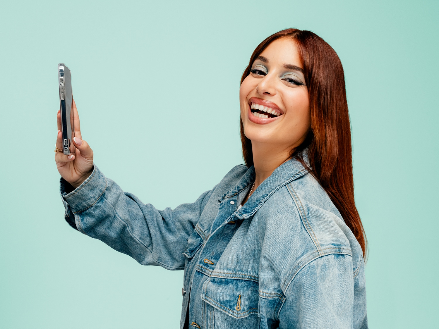 Een lachende vrouw met lang roodbruin haar en een spijkerjasje houdt een zilverkleurige smartphone omhoog tegen een lichtblauwe achtergrond.