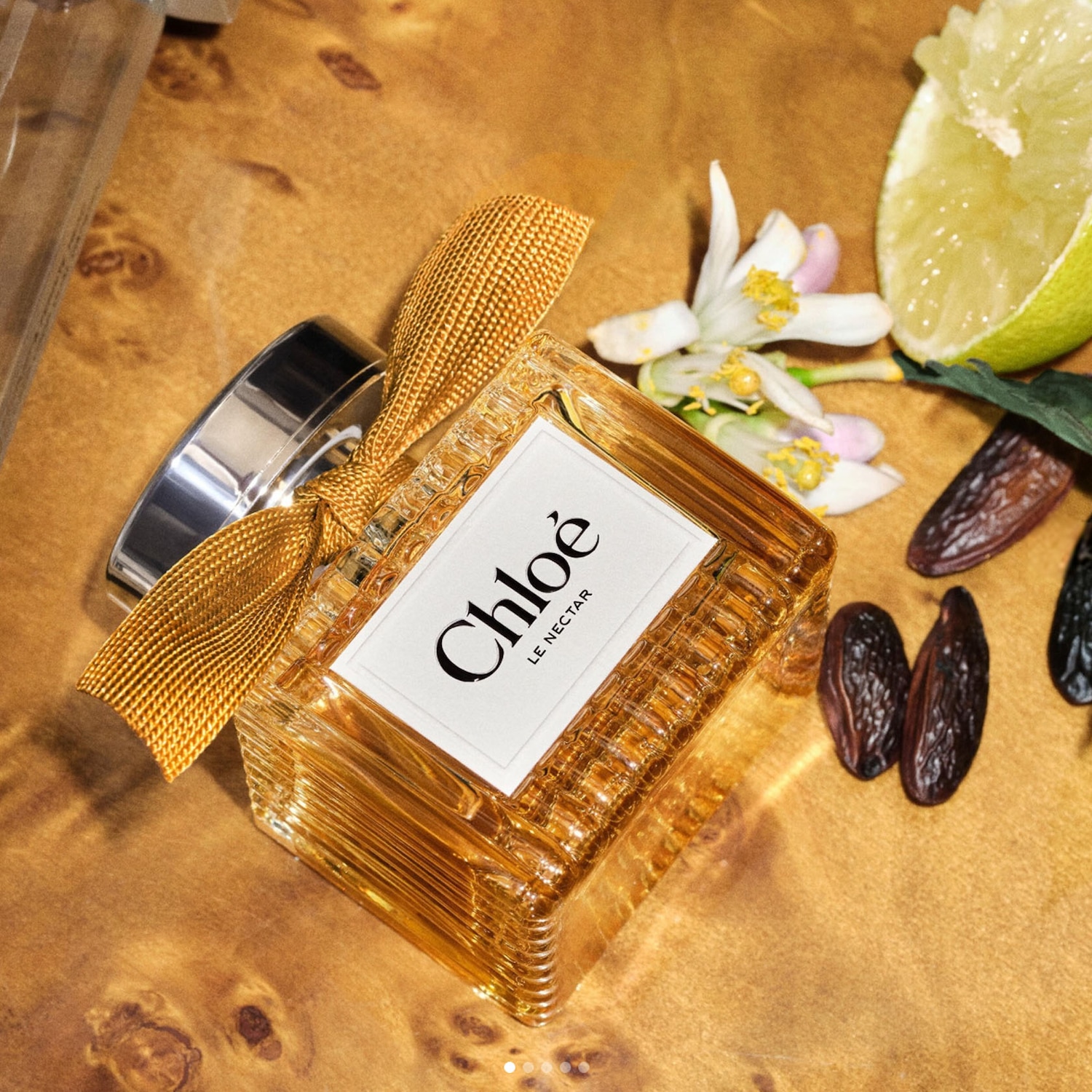 Parfém Chloé Le Nectar ve zlatavé barvě s mašlí, doplněný o citrusy a sušené ovoce, je umístěn na dřevěném podkladu.