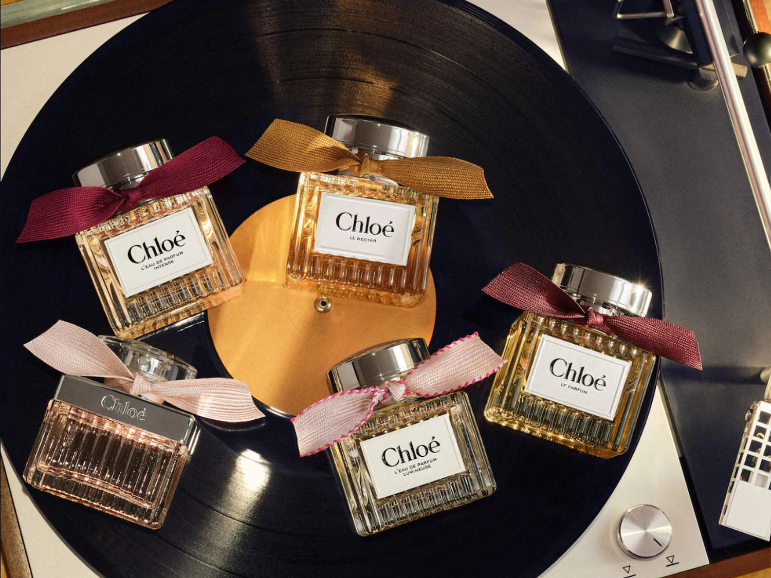 Pět flakonů parfému Chloé s různobarevnými mašlemi, včetně variant L'Eau de Parfum Intense, Le Nectar, L'Eau de Parfum Lumineuse a Le Parfum, je elegantně rozmístěno na černém vinylovém gramofonu.