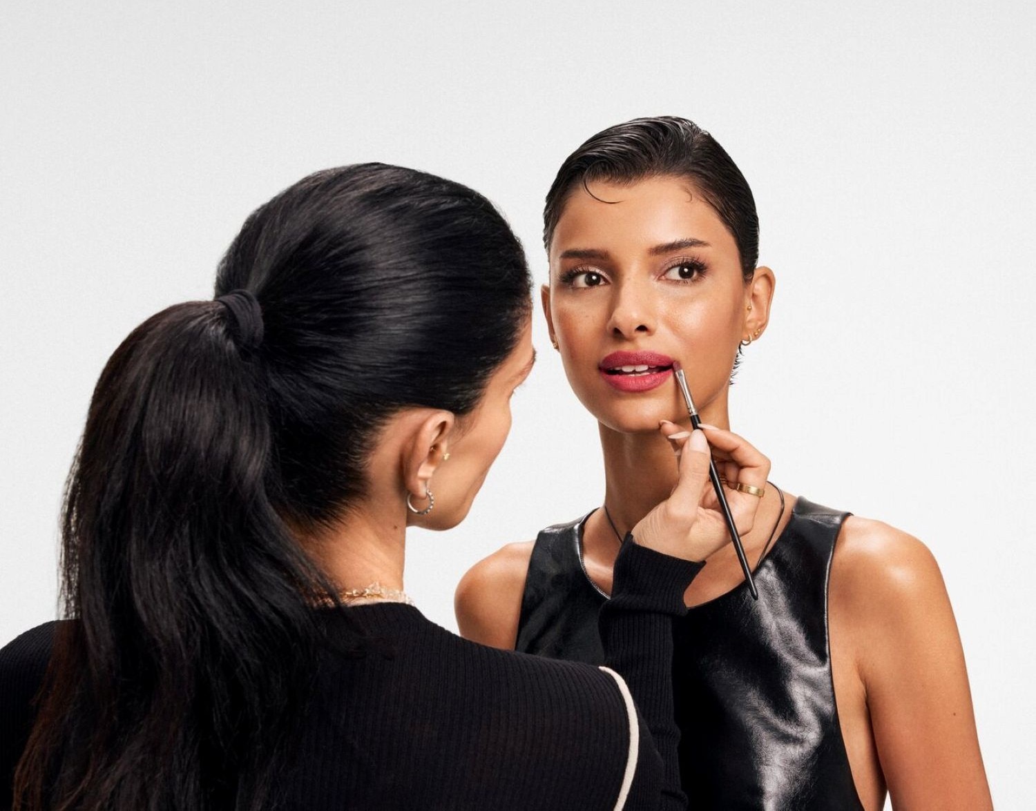 Una truccatrice con i capelli neri legati in una coda di cavallo applica un rossetto rosa brillante sulle labbra di una modella con i capelli scuri tirati all'indietro, che indossa un top nero senza maniche.