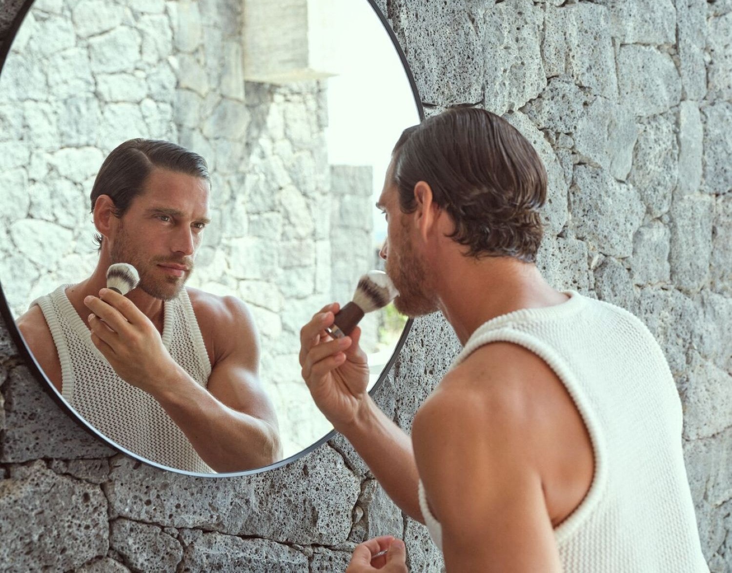 Un uomo con una canotta color crema si guarda allo specchio, tenendo in mano un pennello da barba con setole bianche e marroni, pronto per la rasatura.