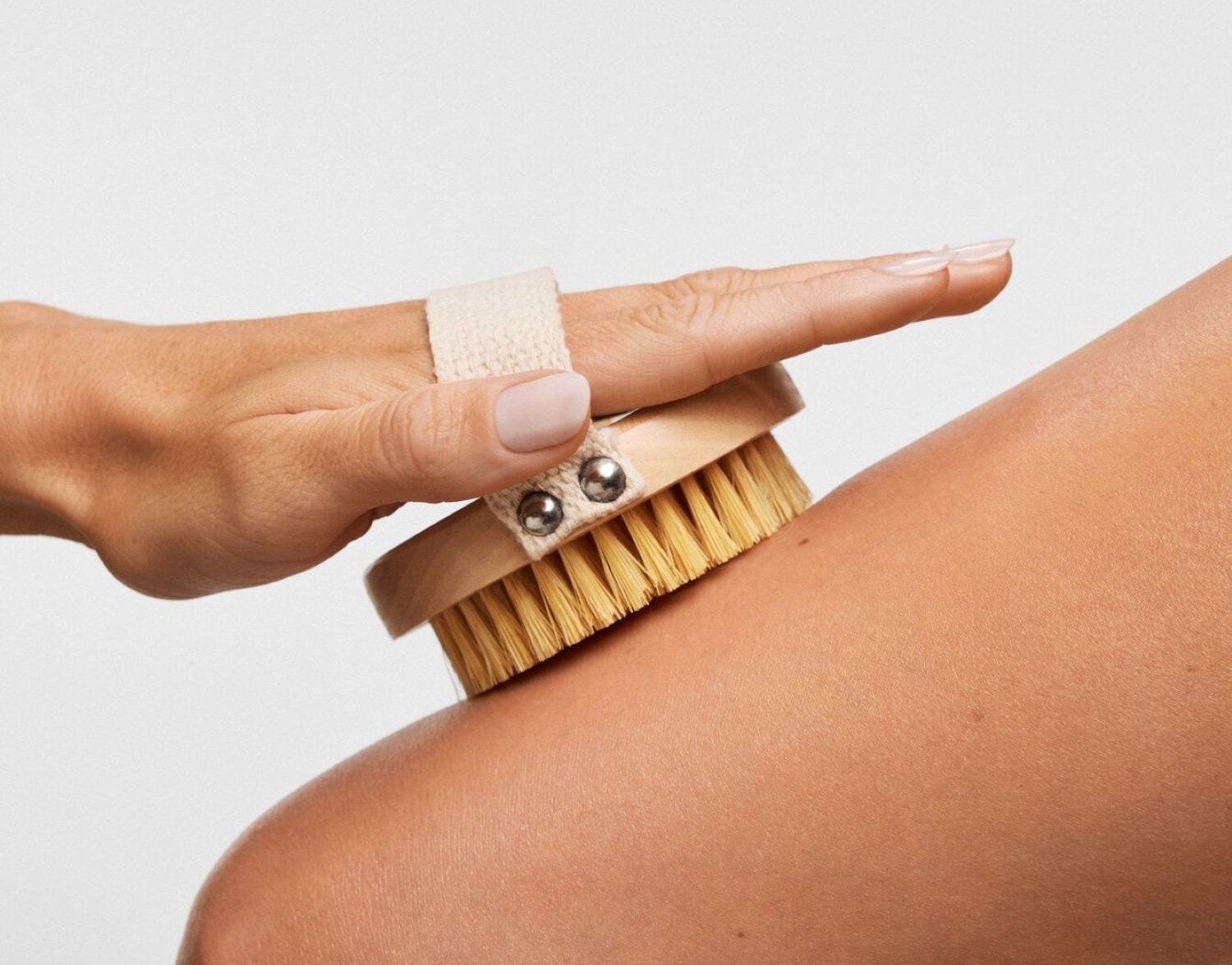 Una mano con unghie curate spazzola delicatamente la pelle del braccio con una spazzola di legno chiara e setole gialle, dotata di un cinturino in tessuto beige per una presa comoda.