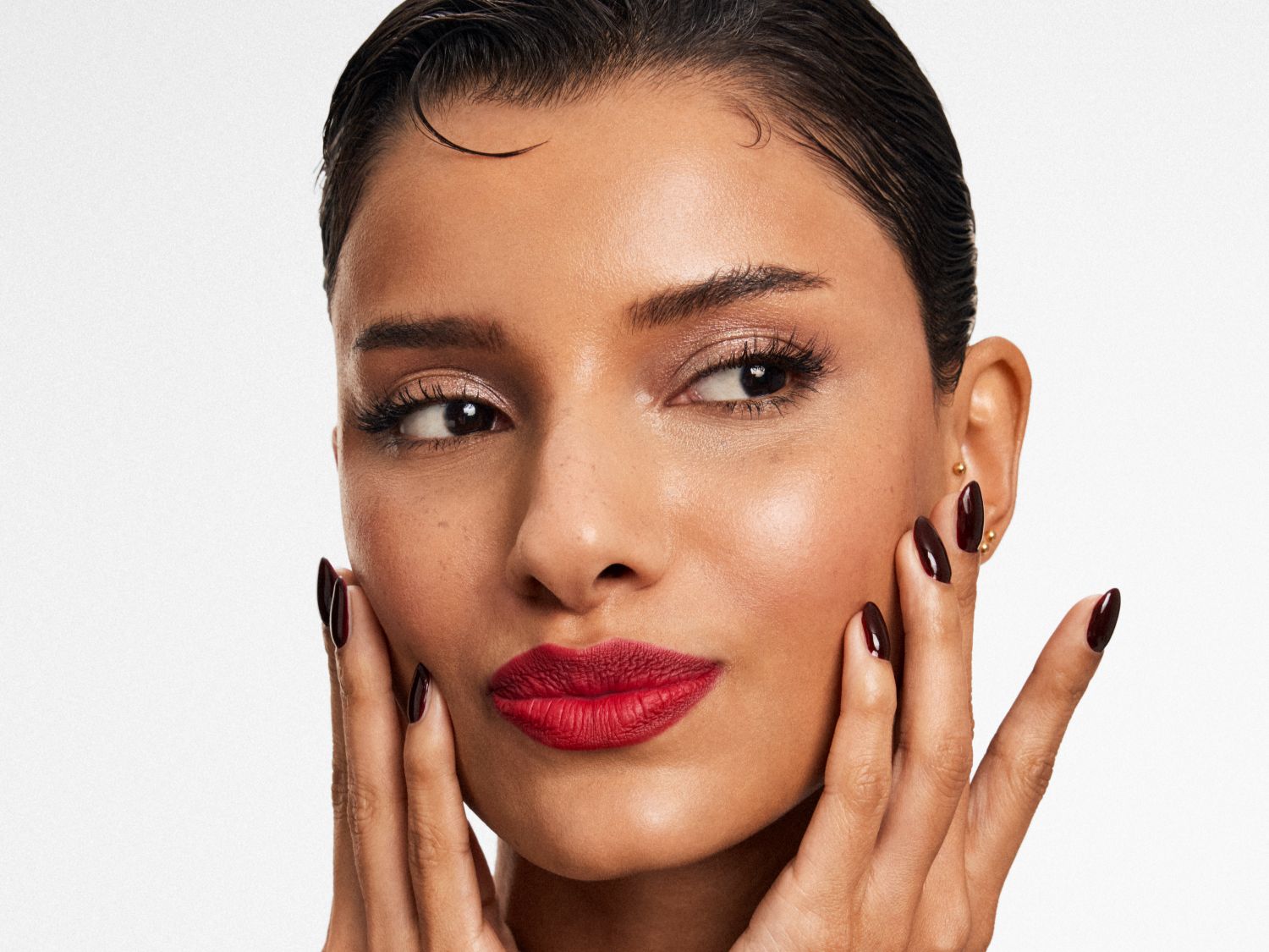Una donna con labbra rosse e unghie bordeaux tocca delicatamente il viso, mettendo in risalto il suo trucco impeccabile e la pelle luminosa.