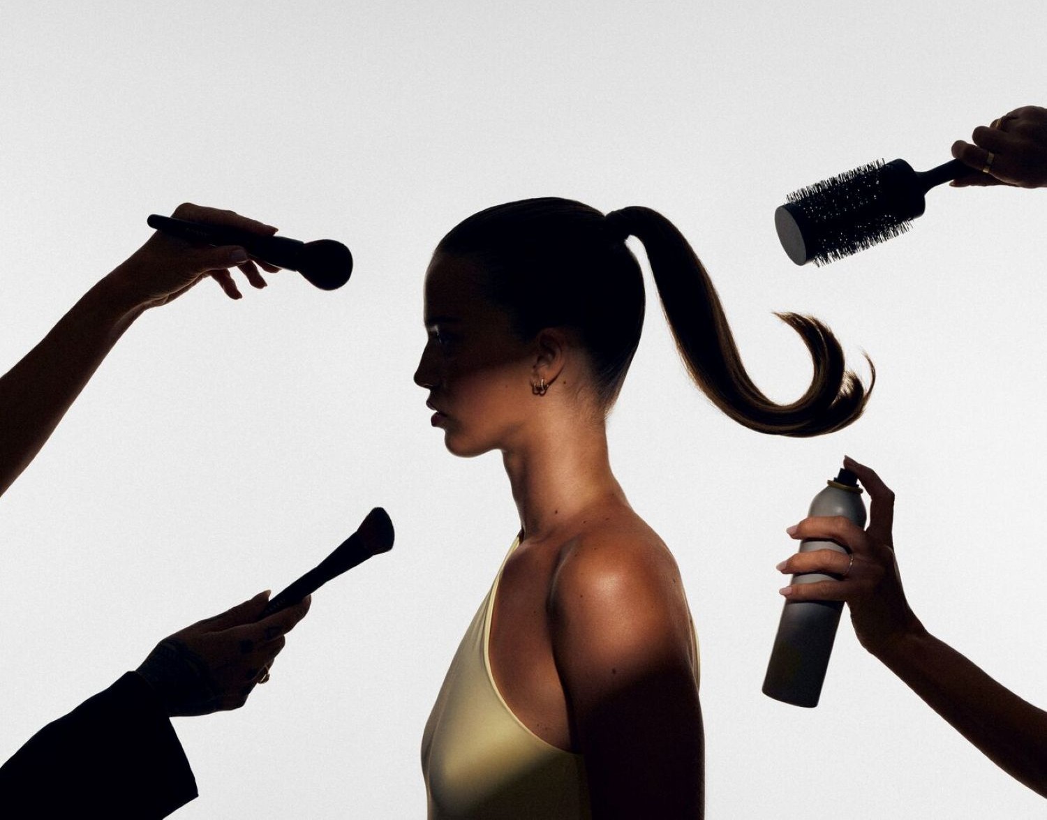 Una donna con una coda di cavallo è circondata da mani che applicano trucco e prodotti per capelli, con pennelli per il viso, una spazzola rotonda e una bomboletta spray per capelli.