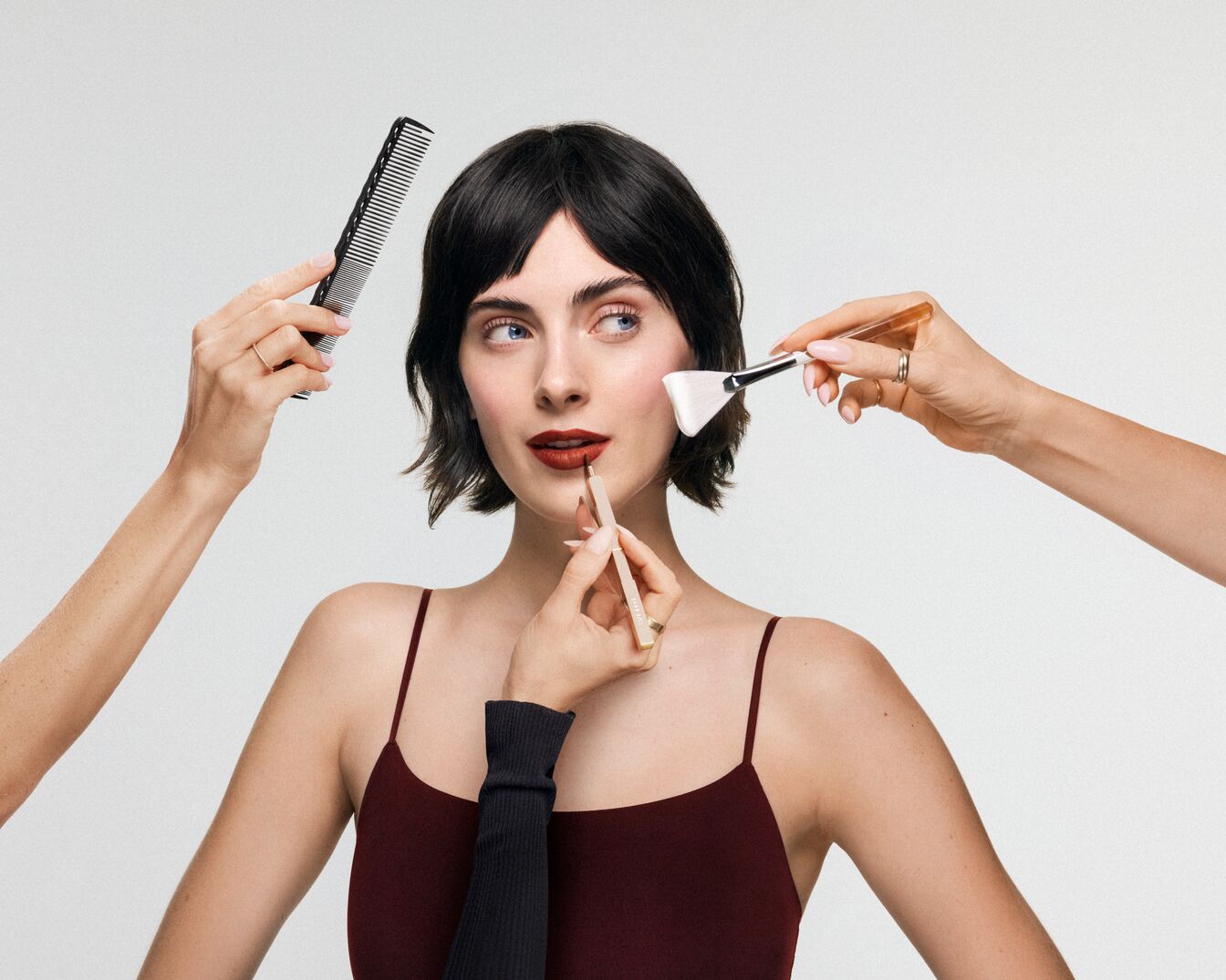Una donna con i capelli corti e scuri, che indossa una canotta bordeaux, si sta truccando le labbra con una matita marrone scuro, mentre diverse mani la aiutano a pettinarsi e ad applicare il trucco.