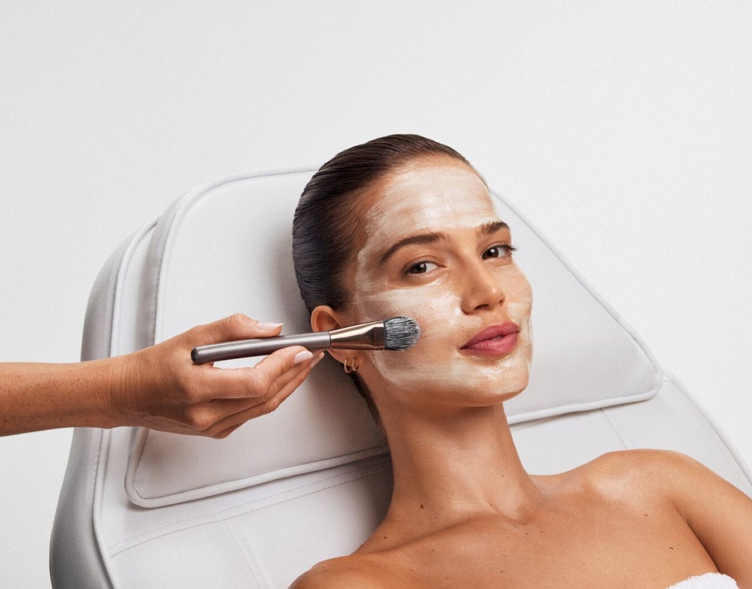 Una donna con una maschera bianca sul viso, applicata con un pennello da una mano, è sdraiata su una sedia bianca, con i capelli tirati indietro e le labbra rosa.