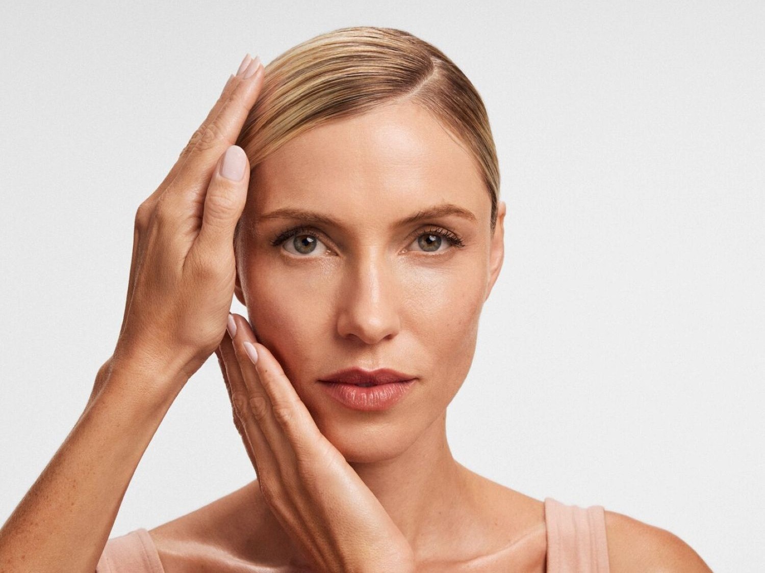 Una donna bionda con la pelle chiara e luminosa tocca delicatamente il viso con entrambe le mani, mettendo in risalto la sua carnagione impeccabile e i suoi occhi chiari.