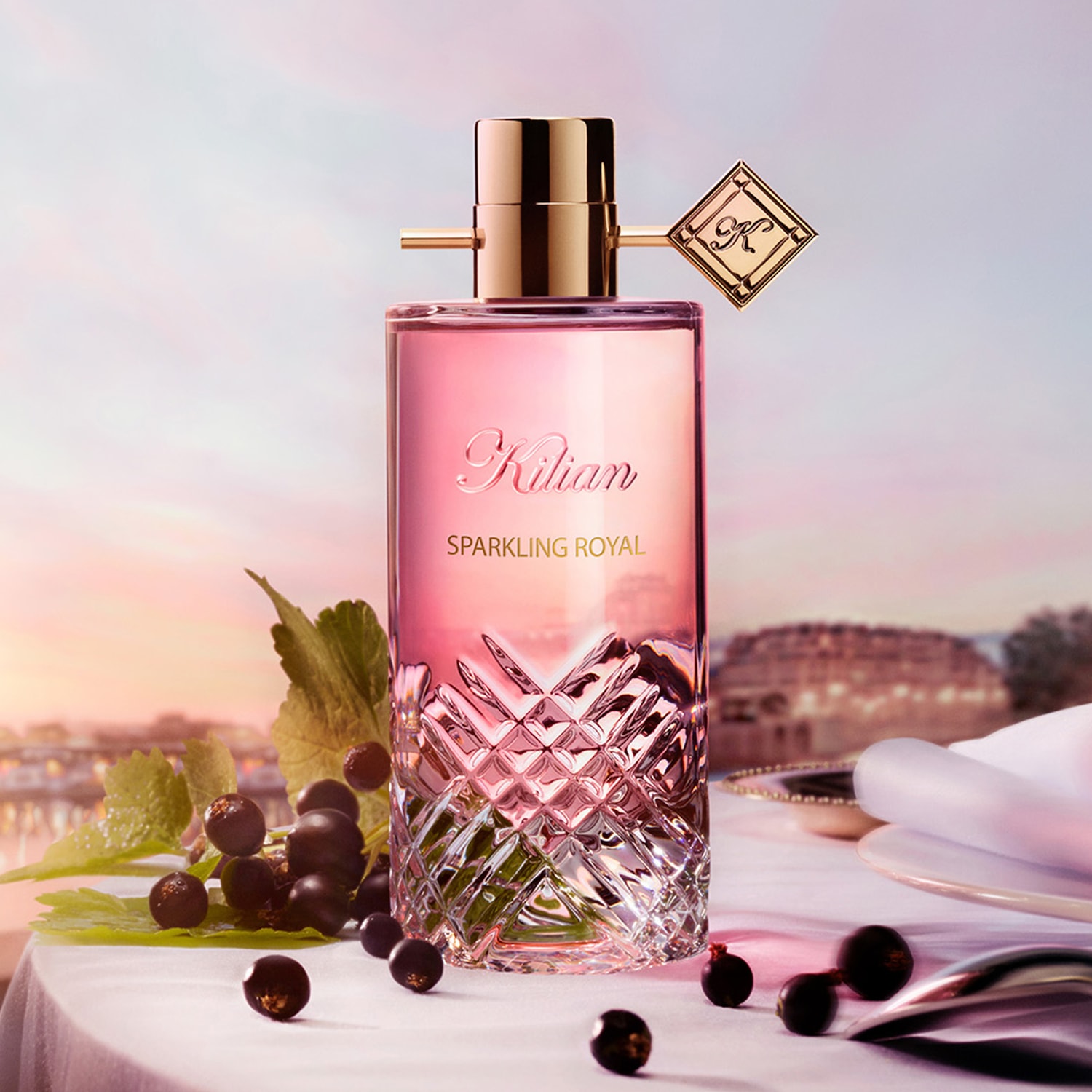 Sparkling Royal präsentiert sich in einem eleganten, transparenten Glasflakon mit goldenem Verschluss und einem dekorativen goldenen Anhänger, umgeben von Schwarzen Johannisbeeren auf einem festlich gedeckten Tisch.