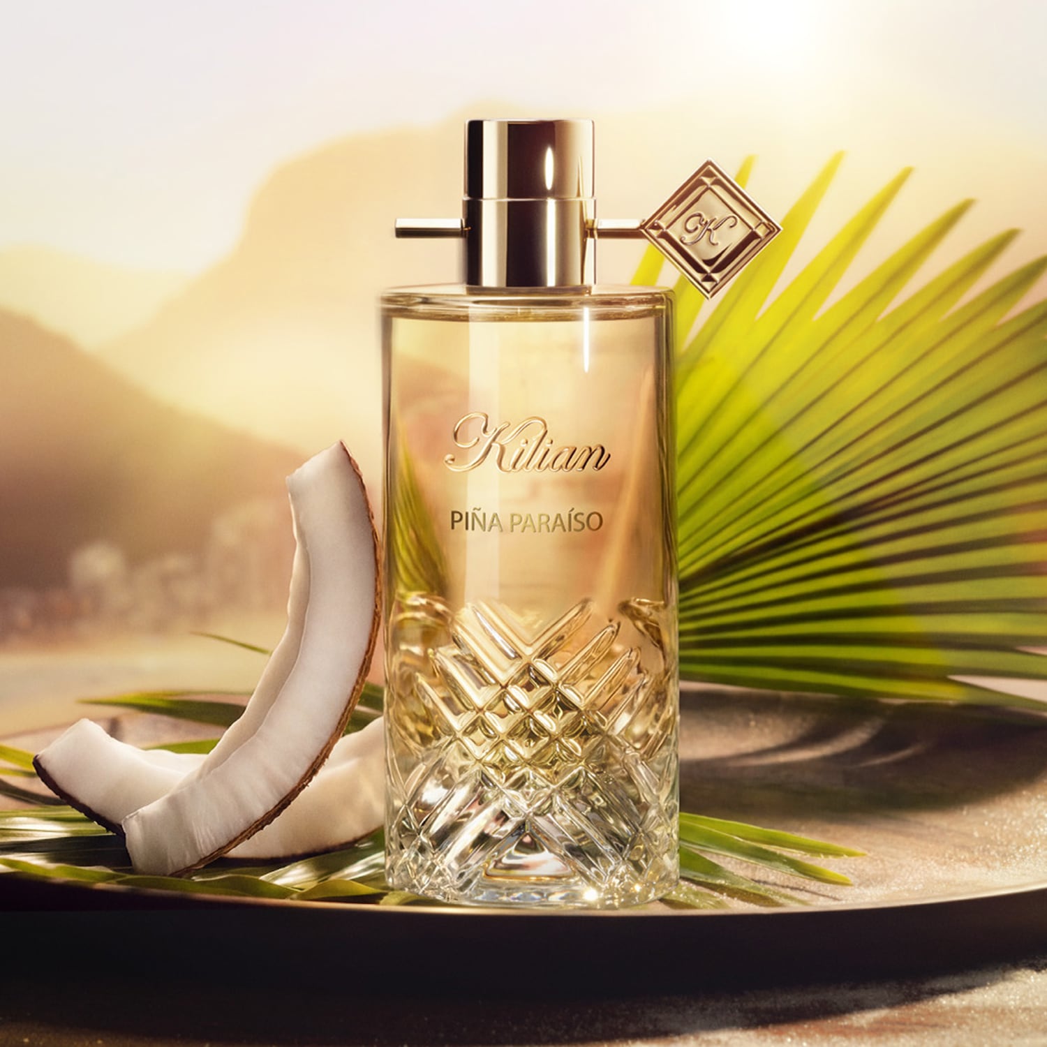 Piña Paraíso in einem eleganten, facettierten Glasflakon mit goldenem Verschluss und Anhänger, umgeben von Kokosnussstücken und Palmblättern auf einem sandigen Tablett.