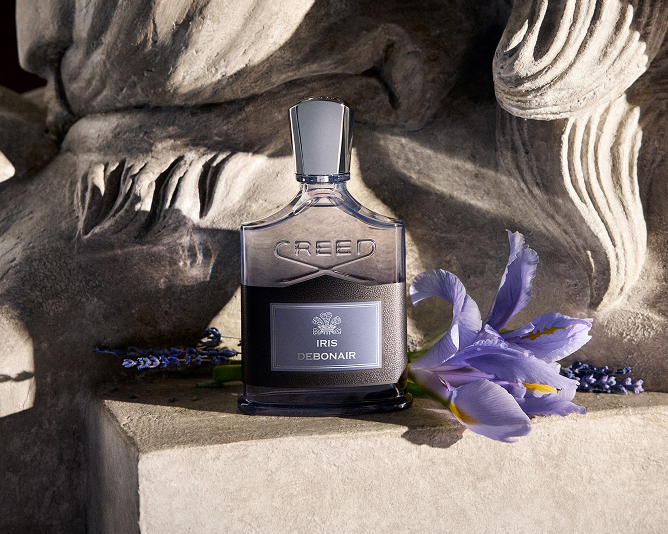 Parfém Creed Iris Debonair v hranaté lahvičce s černým koženým obalem a stříbrným uzávěrem, doplněný fialovým květem kosatce a levandulí, je umístěn na kamenném podstavci s vyřezávanými detaily.
