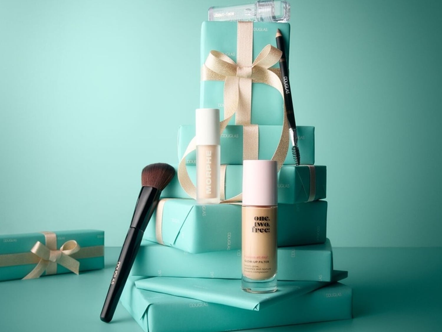 Un set de machiaj festiv, cu produse cosmetice așezate pe cutii de cadouri turcoaz, legate cu panglici aurii, include o pensulă de machiaj neagră, un fond de ten lichid One.Two.Free! Glow-Up Filter, un corector Morphe și un creion de sprâncene Douglas.