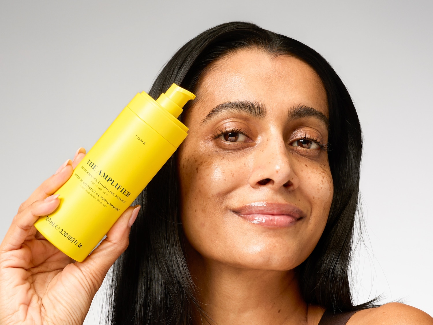 A woman with dark hair and glowing skin holds a bright yellow bottle of "The Amplifier Performance Enhancing Essence" by TONE, designed for all skin types, containing 100 mL.
