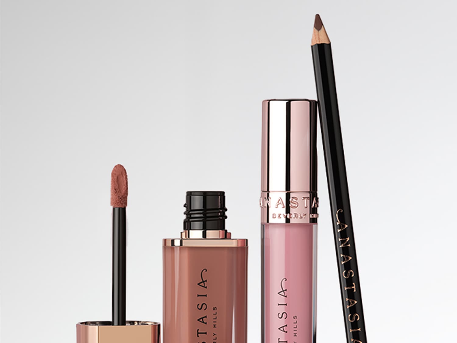 Drei Anastasia Beverly Hills Lippenprodukte stehen nebeneinander vor hellem Hintergrund: ein matter flüssiger Lippenstift in Nude-Braun, ein glänzender Lippenstift in hellem Rosa und ein brauner Lipliner.