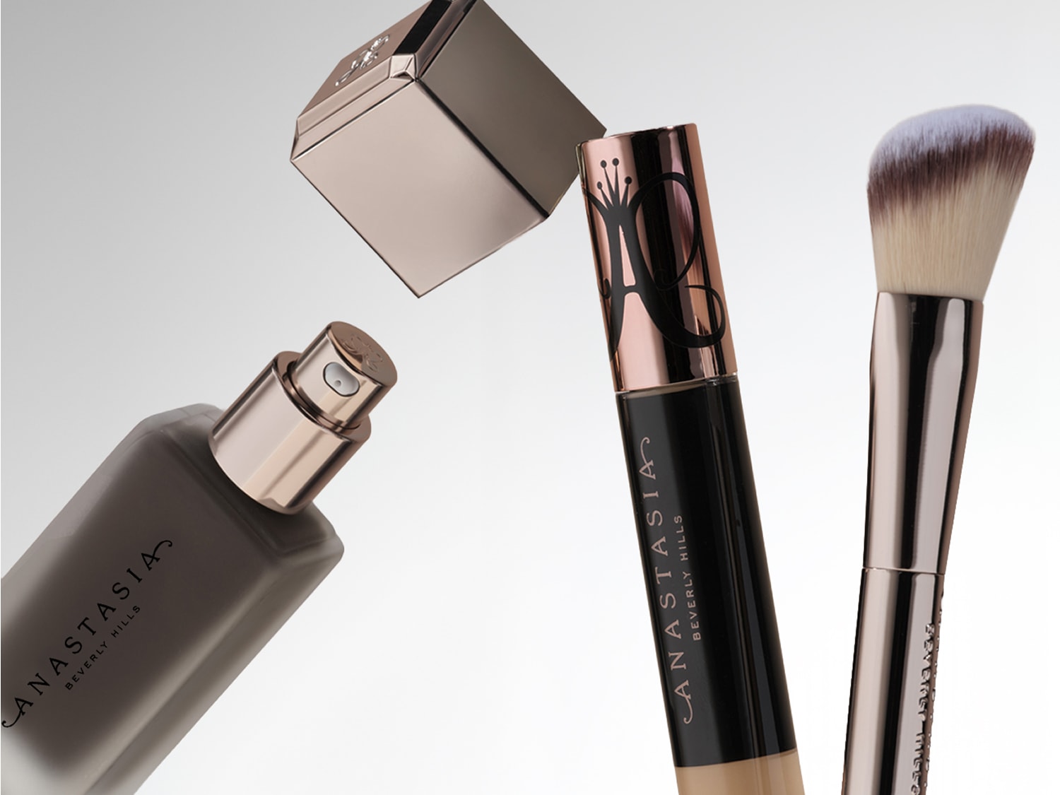 Das Bild zeigt drei Anastasia Beverly Hills Produkte: eine Foundation in einem matten Glasflakon mit geöffneter, goldfarbener Kappe, einen Concealer in einem schwarzen Flakon mit goldener Kappe und einen Make-up-Pinsel mit goldenem Griff und weißen Borsten.