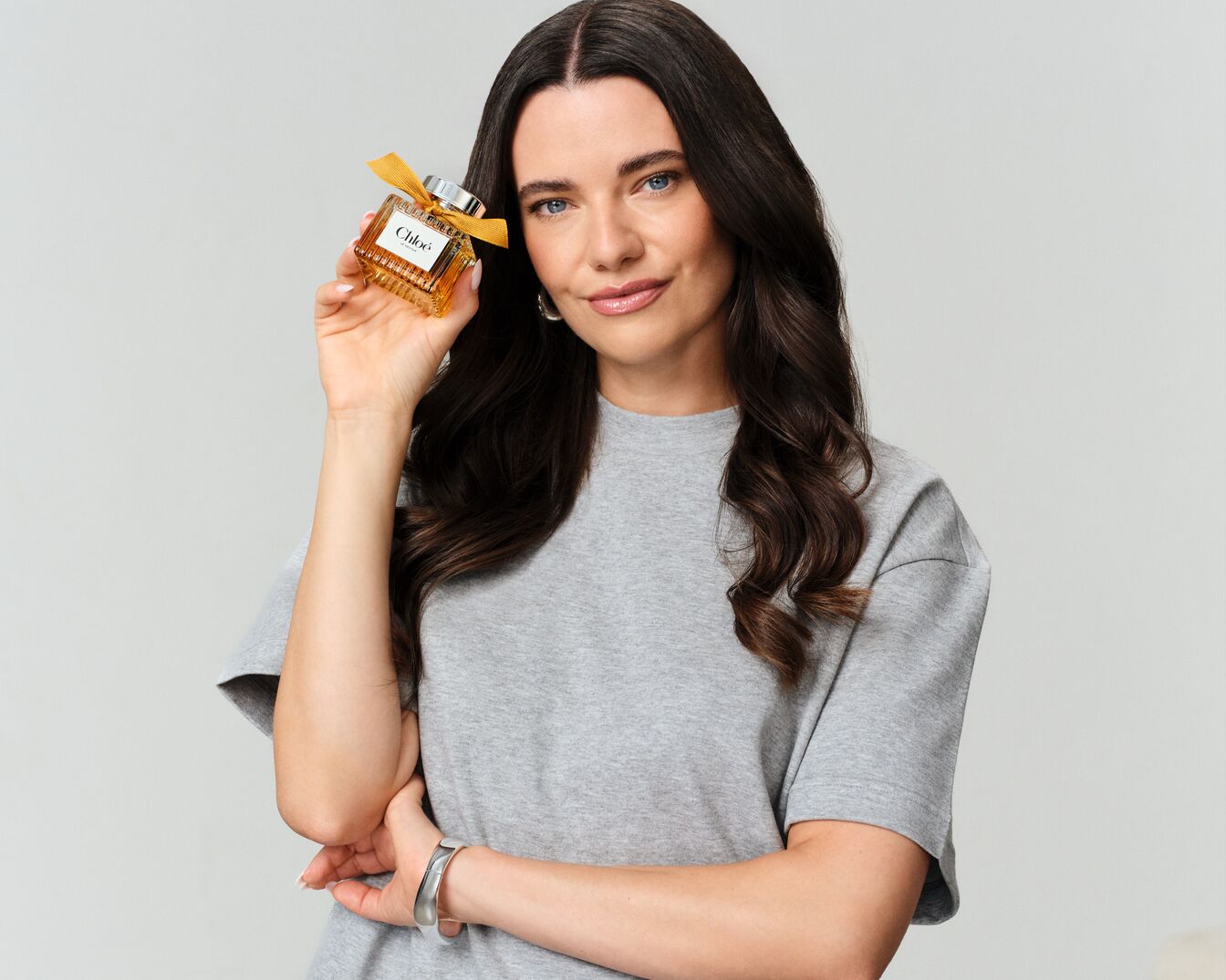 Eine Frau mit langen, dunkelbraunen Haaren und einem grauen T-Shirt hält einen quadratischen Chloé-Duftflakon mit goldener Flüssigkeit und einer gelben Schleife in der Hand.
