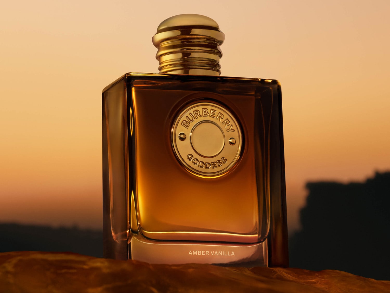 Parfém Burberry Goddess Amber Vanilla v obdélníkové lahvičce s jantarovým odstínem a zlatým uzávěrem, s kulatým zlatým štítkem s nápisem „Burberry Goddess“ uprostřed, stojí na texturovaném povrchu s rozmazaným oranžovým pozadím.