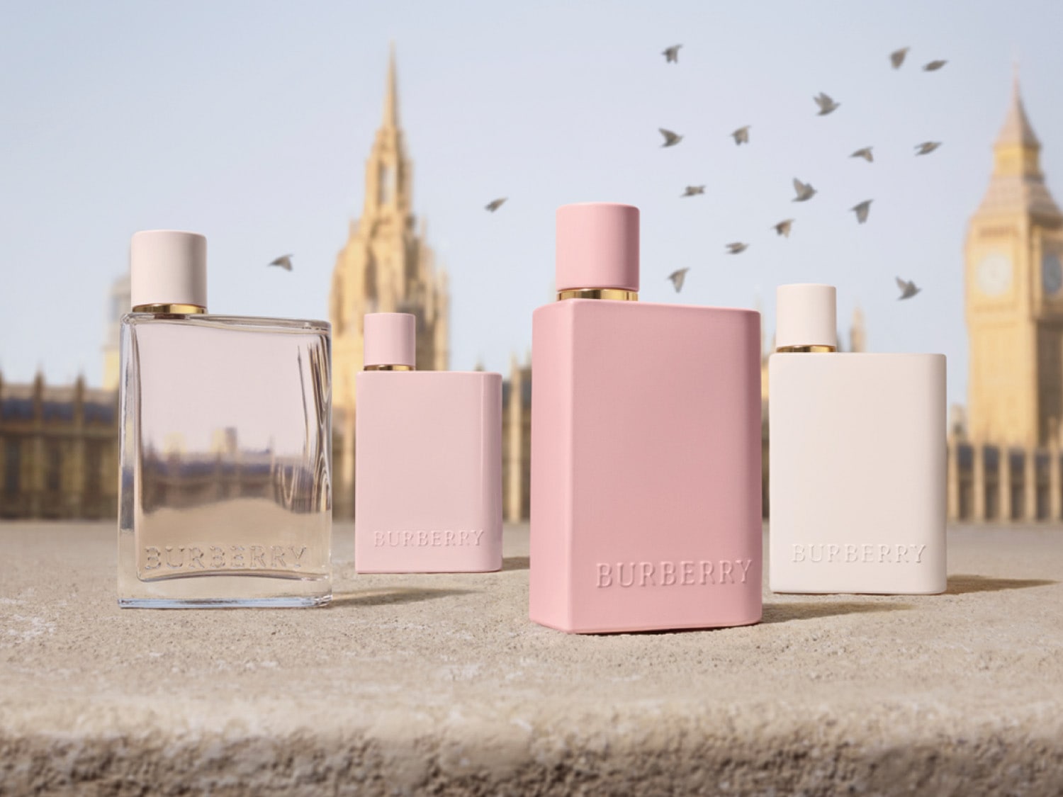 Čtyři flakony parfému Burberry, dva průhledné a dva neprůhledné v růžových a krémových odstínech, stojí na kamenném podkladu s rozmazaným pozadím londýnských památek a ptáků letících po obloze.