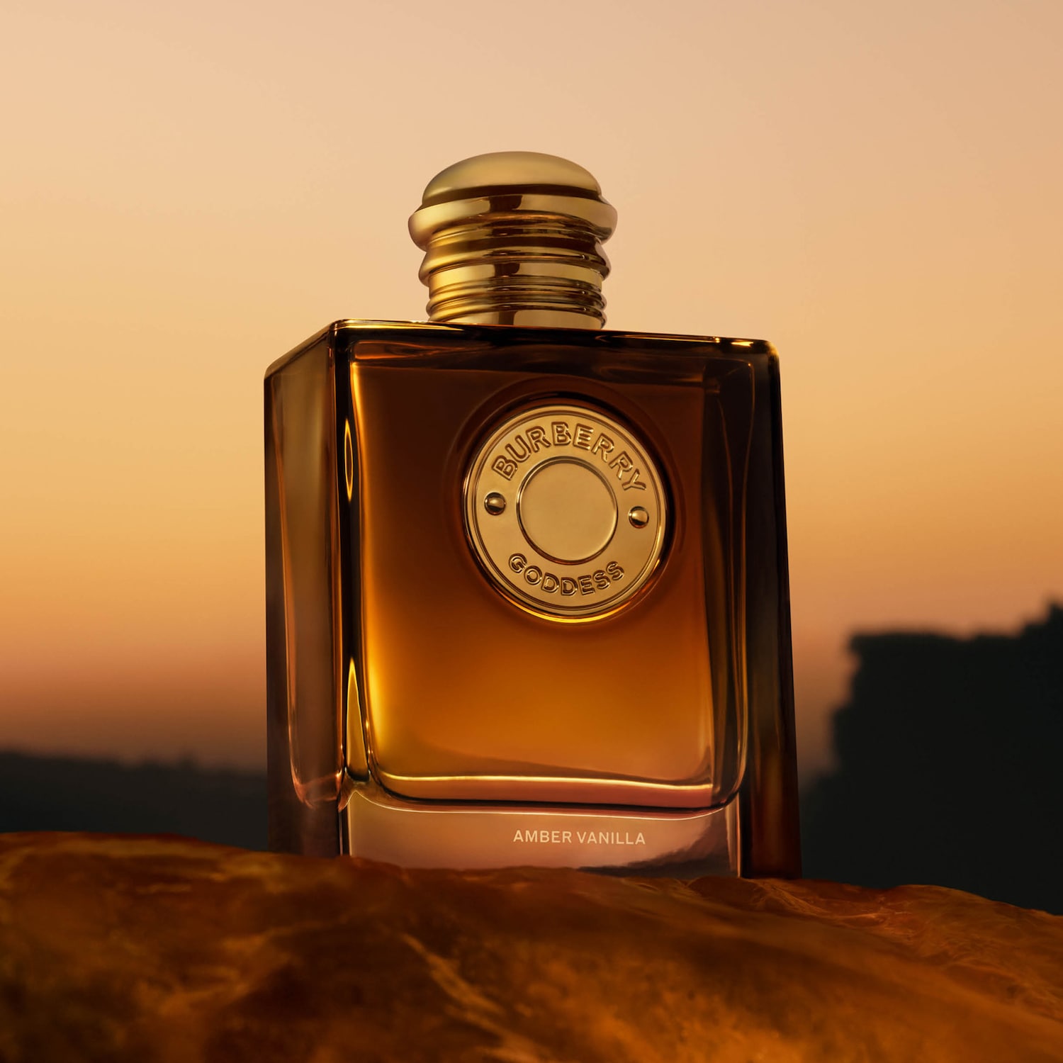Parfém Burberry Goddess Amber Vanilla v obdélníkovém flakonu s jantarovou tekutinou a zlatým uzávěrem, s kulatým zlatým štítkem s nápisem "BURBERRY GODDESS" uprostřed, stojí na hnědém podkladu s rozmazaným oranžovým pozadím.
