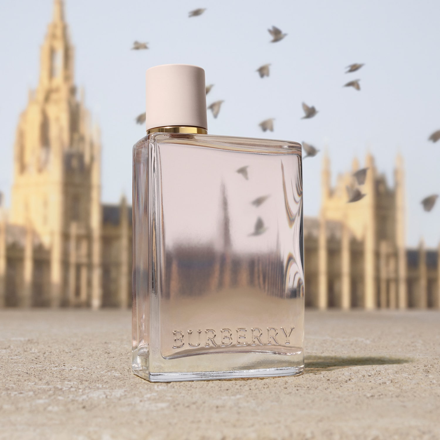 Parfém Burberry Her London Dream v obdélníkové skleněné lahvičce s béžovým uzávěrem a zlatým lemem, s nápisem "BURBERRY" na přední straně, stojí na pískovém povrchu s rozmazaným pozadím budov a ptáků.
