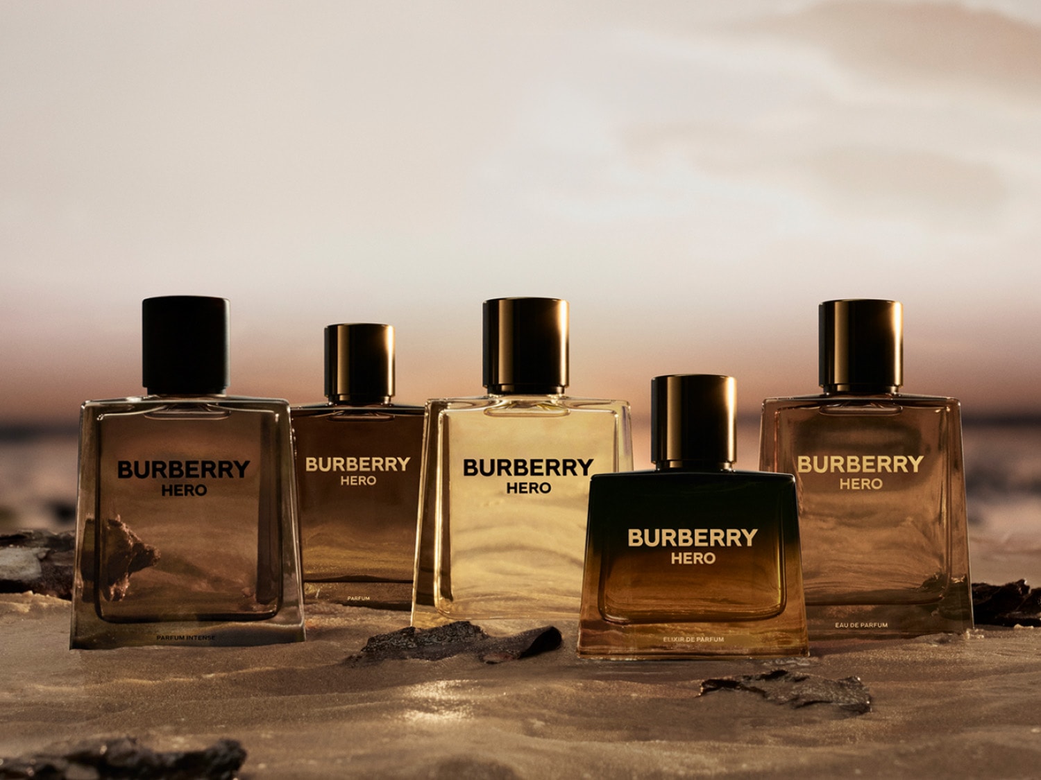 Pět lahviček parfému Burberry Hero s černými uzávěry a různými odstíny jantarové tekutiny, od světlé po tmavou, stojí na písečném povrchu s rozmazaným pozadím západu slunce.