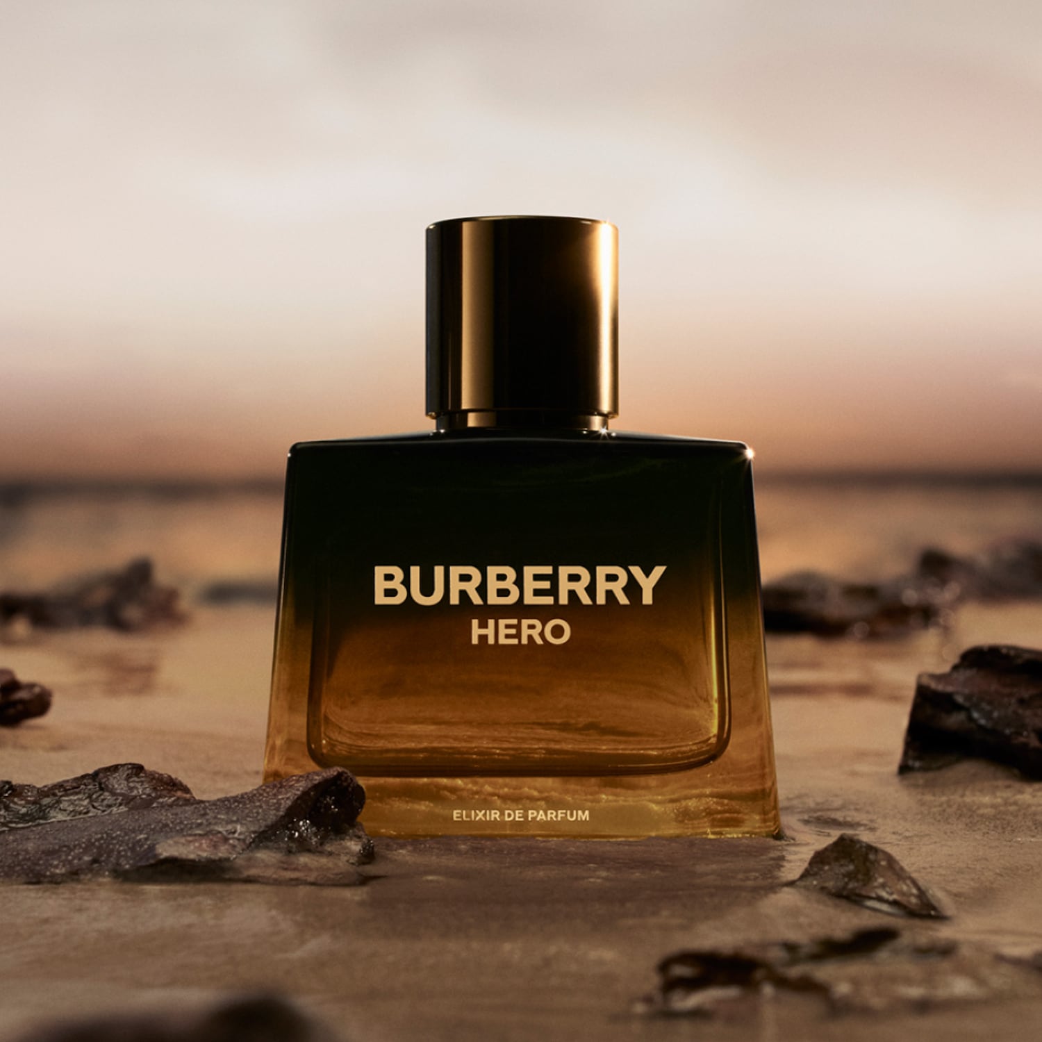 Parfém Burberry Hero Elixir de Parfum v tmavě jantarové lahvičce se zlatým uzávěrem stojí na mokrém písku s kameny, s rozmazaným pozadím západu slunce.