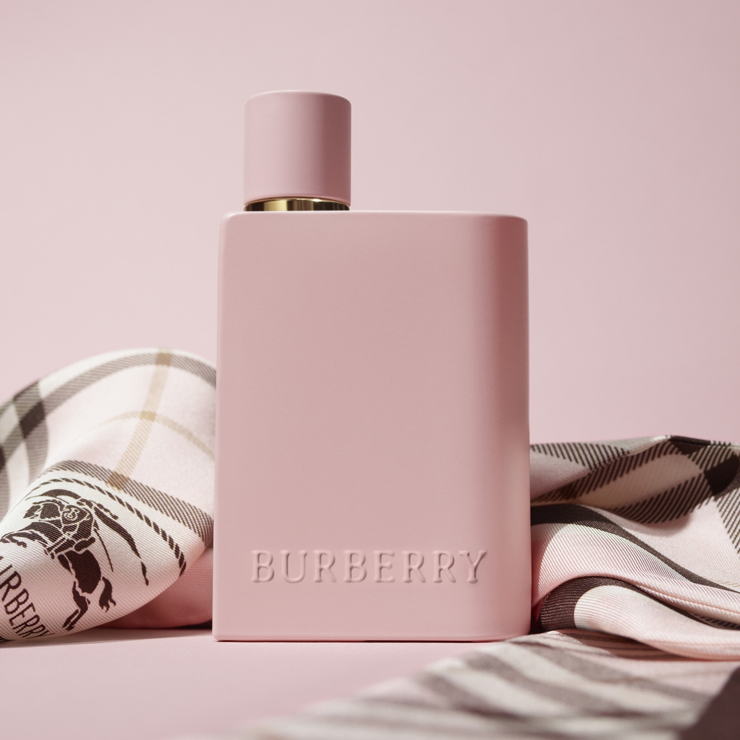 Růžový flakon parfému Burberry s vyraženým logem značky, doplněný hedvábným šátkem s kostkovaným vzorem, leží na světle růžovém pozadí.