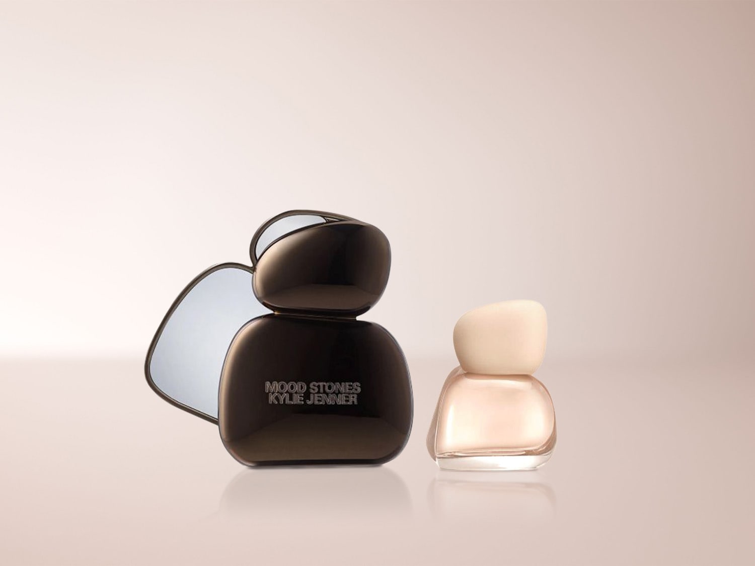 Zwei Nagellackflaschen von Kylie Jenner, eine in glänzendem Schwarz mit der Aufschrift "MOOD STONES KYLIE JENNER" und eine kleinere in hellem Rosé, stehen nebeneinander vor einem Spiegel.