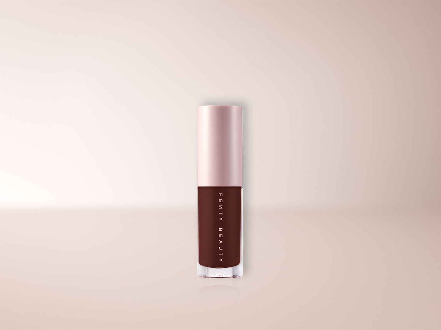 Un tube de brillant à lèvres Fenty Beauty marron foncé avec un capuchon rose pâle est présenté sur un fond rose clair, le nom de la marque est écrit verticalement en blanc sur le tube.