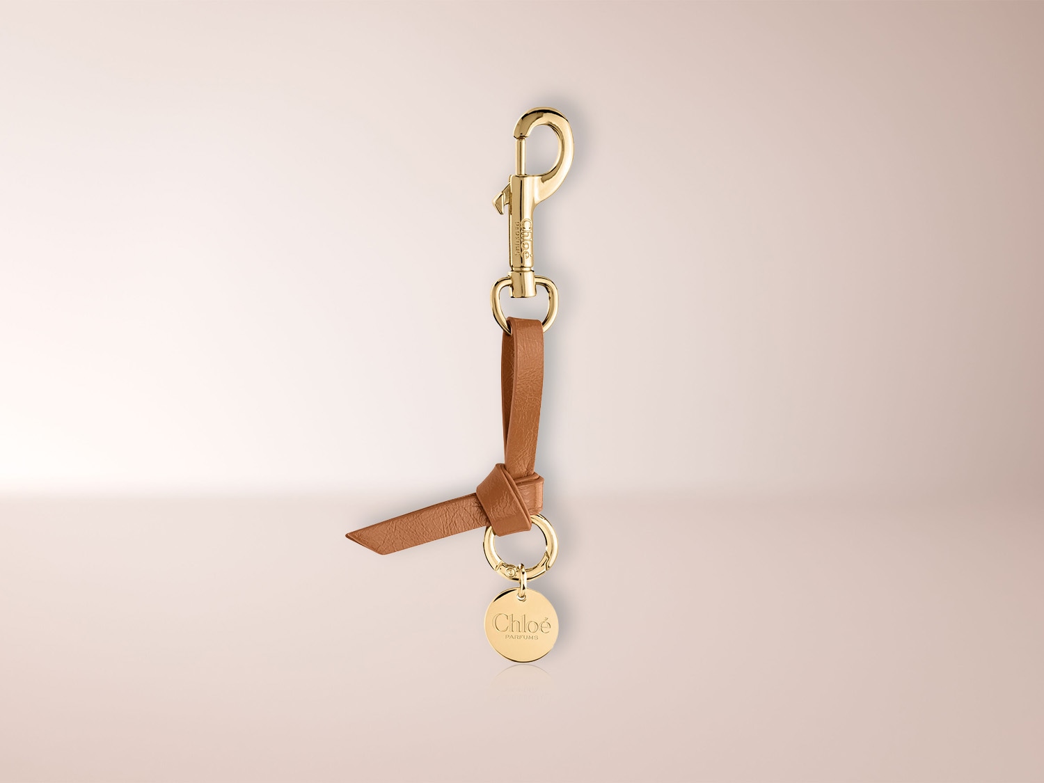 Un porte-clés Chloé en cuir marron clair avec un fermoir mousqueton doré et un médaillon rond doré gravé du logo Chloé Parfums.