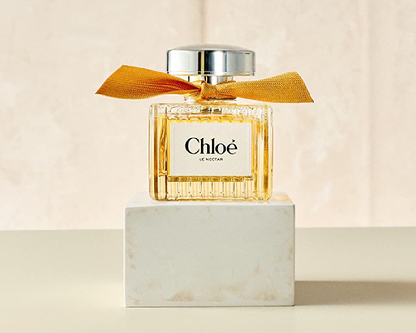 Der Chloé Le Nectar Duft ist in einem quadratischen Glasflakon mit geriffelten Seiten und einem goldenen Band um den Hals verpackt, der auf einem weißen Marmorblock steht.