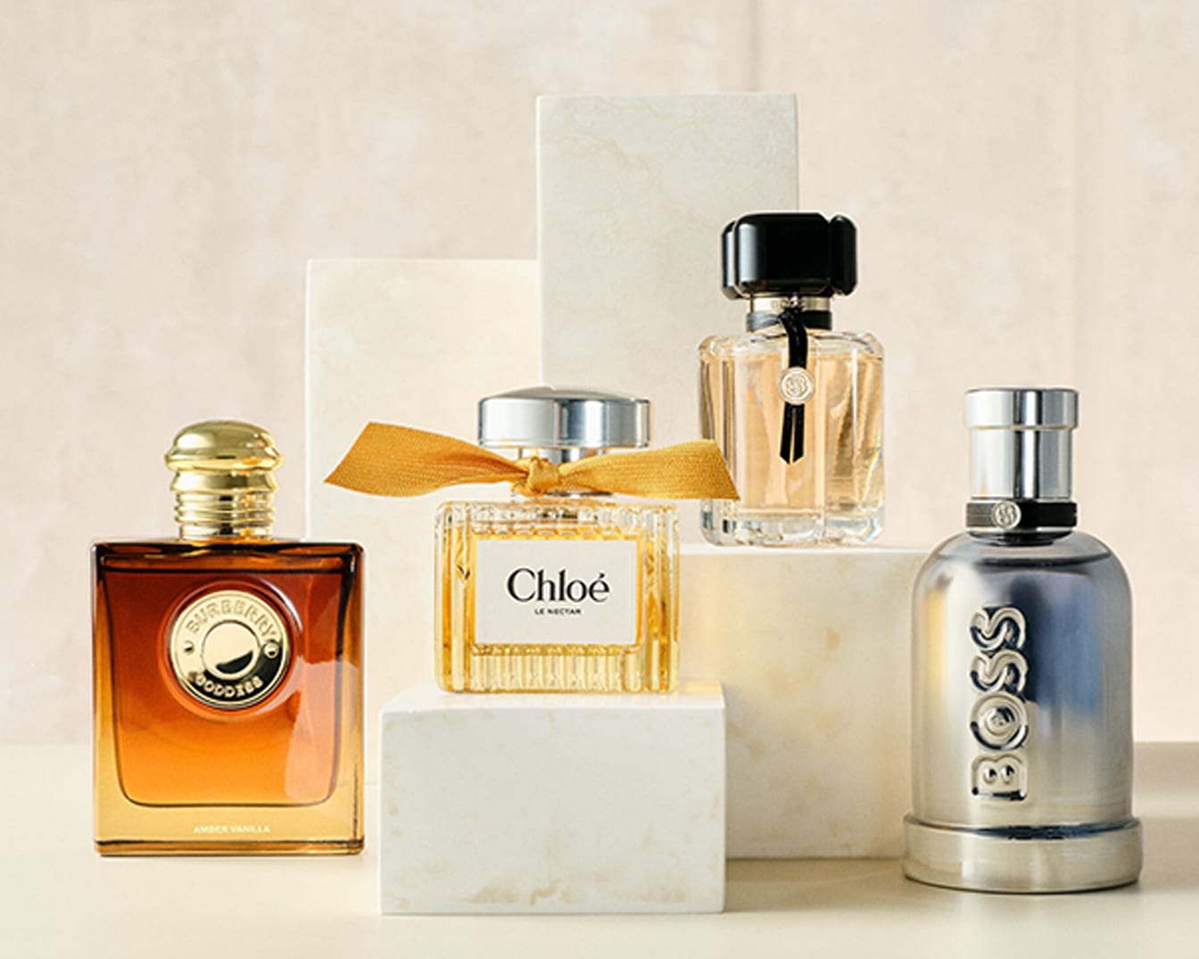 Vier Parfümflaschen, darunter Burberry Goddess Amber Vanilla, Chloé Le Nectar, ein Parfüm mit schwarzer Schleife und ein silberner BOSS-Flakon, stehen auf beigefarbenen Blöcken vor einem hellen Hintergrund.