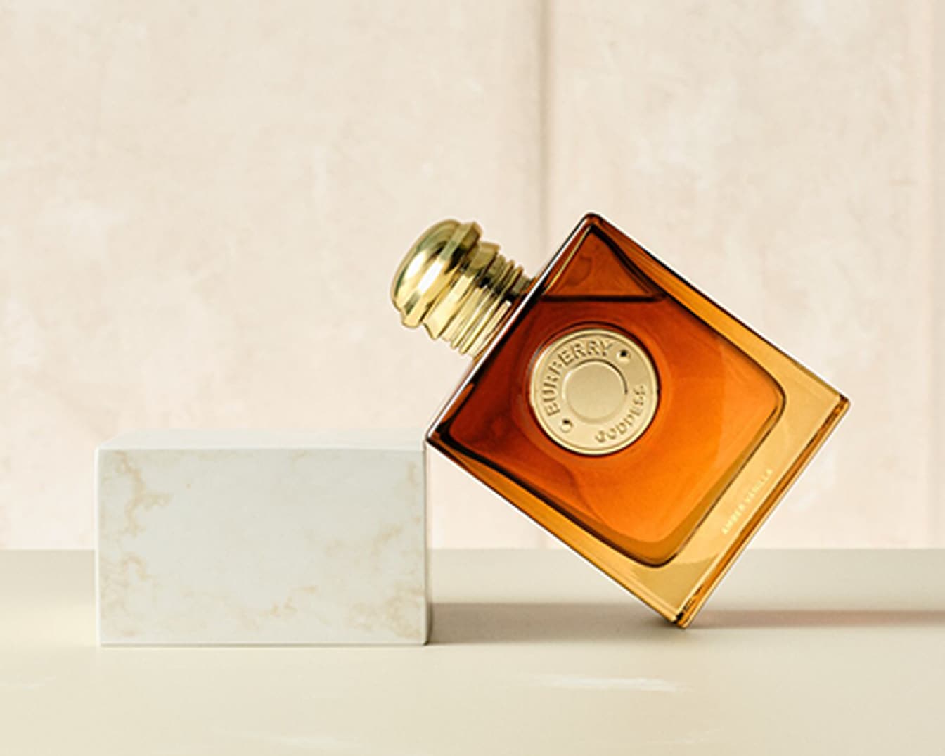 Ein schräg stehender, quadratischer Burberry Goddess-Parfümflakon mit goldener Kappe und bernsteinfarbener Flüssigkeit, der auf einem weißen Marmorblock balanciert.