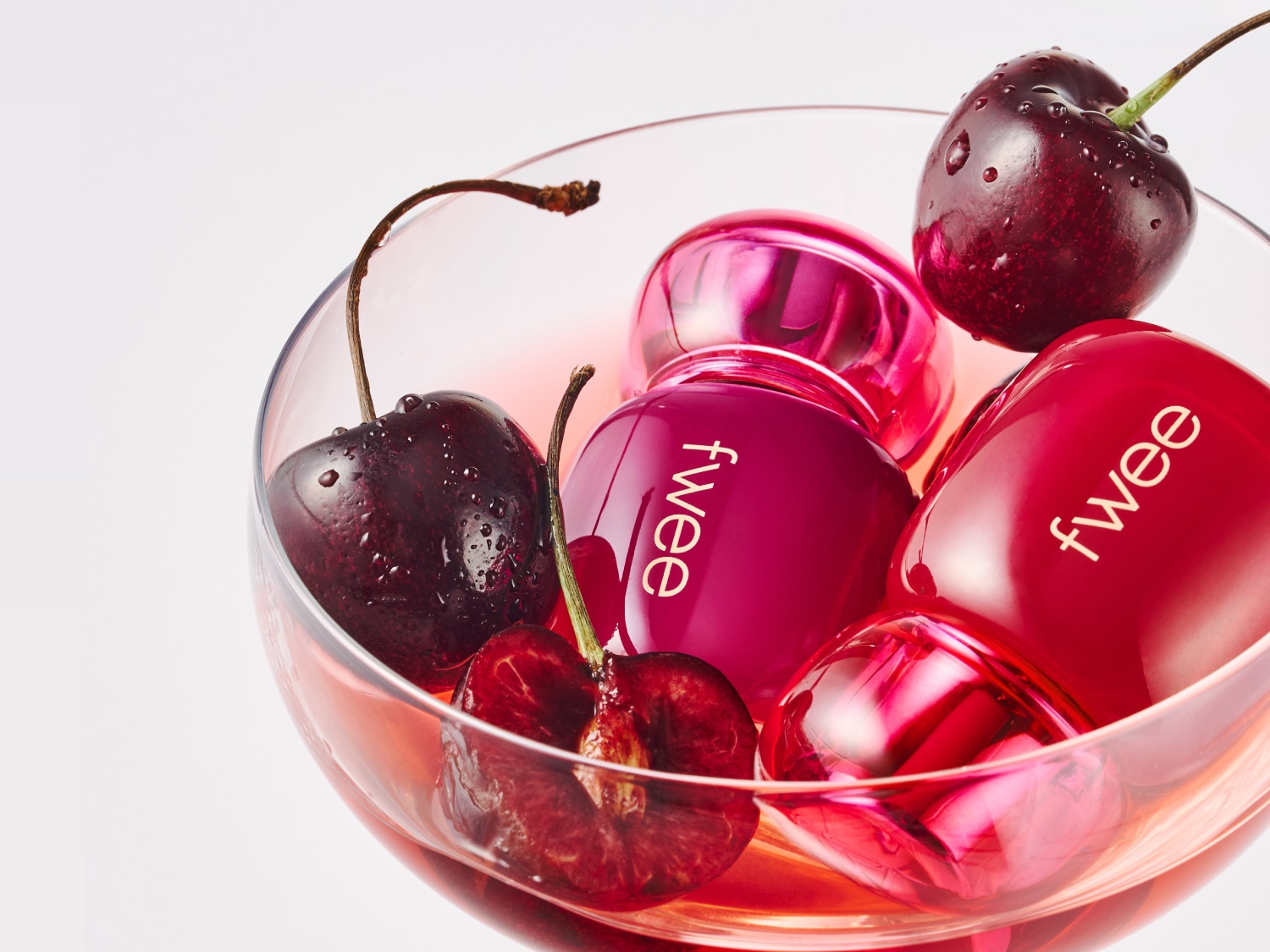 Un verre à cocktail transparent contient des cerises rouges et des flacons de parfum Fwee rouges et roses, créant une ambiance fruitée et élégante.